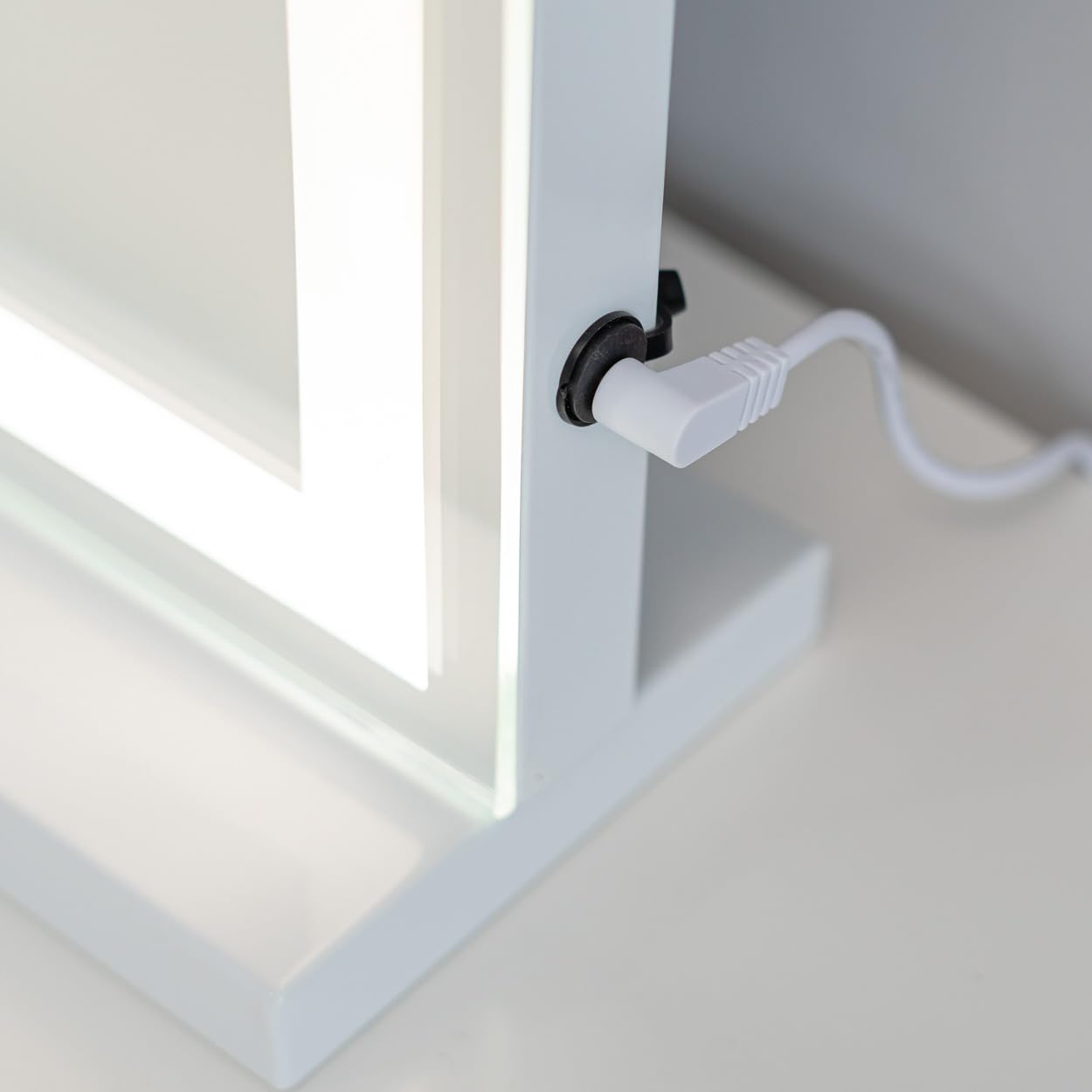 Un espejo de maquillaje blanco iluminado con un cable de alimentación enchufado. El espejo está sobre una superficie blanca.
