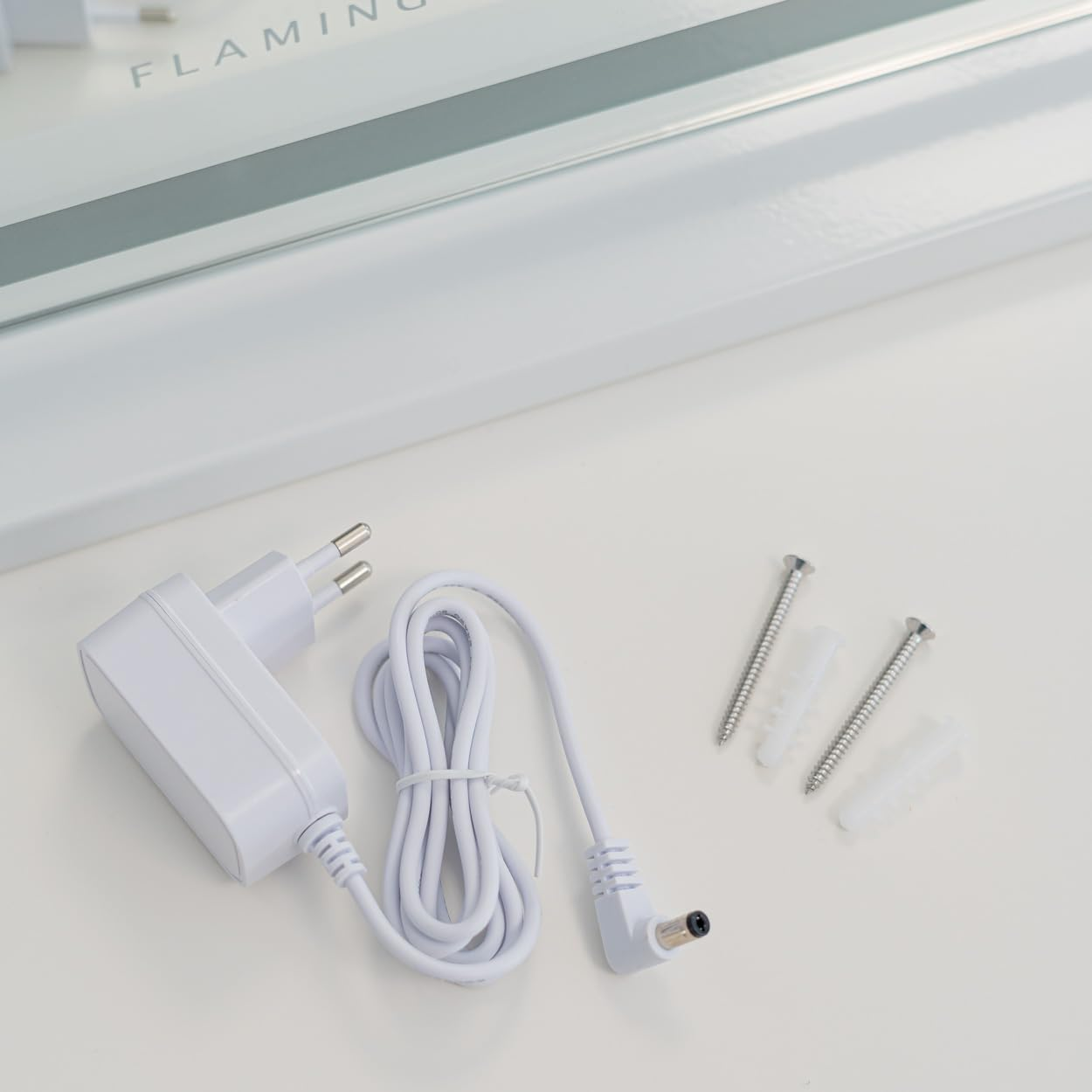Adaptador de corriente blanco, cable, tornillos y anclajes en superficie blanca. Parte de una lámpara al fondo.