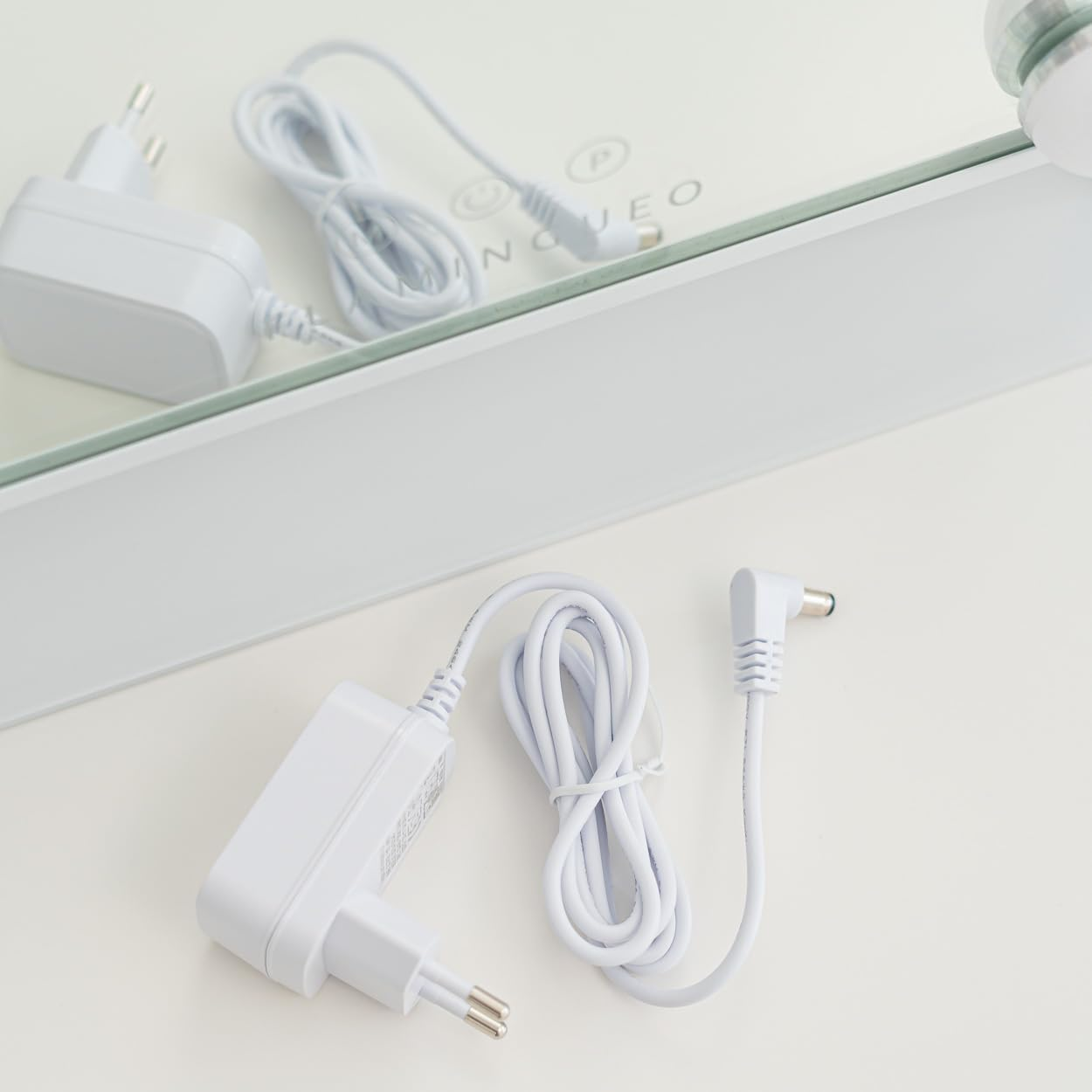 Adaptador de corriente y cable blancos sobre una superficie blanca. Reflejo de un espejo y otro adaptador en el fondo.