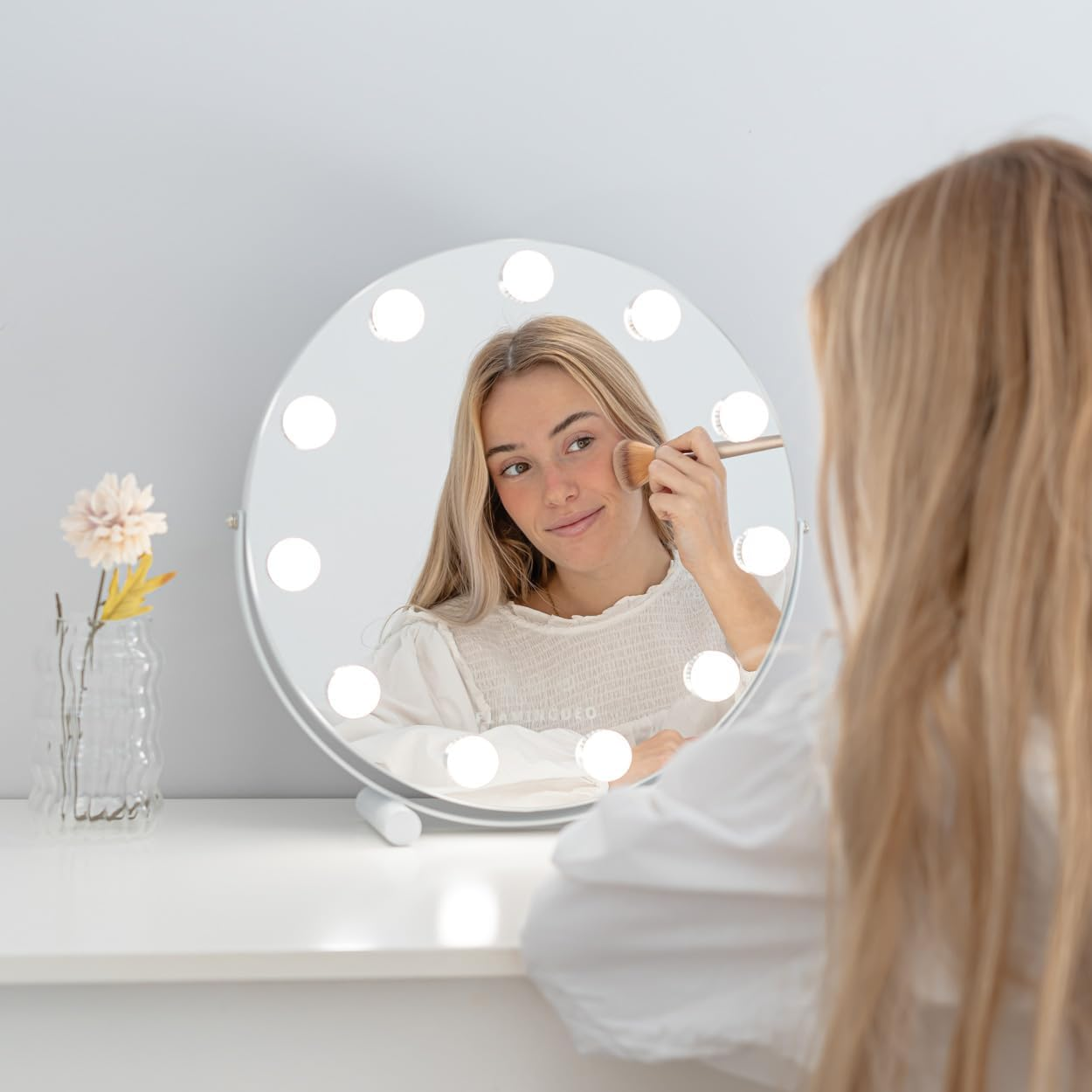 Mujer aplicándose maquillaje con brocha, reflejada en espejo iluminado. Flor y jarrón sobre mesa blanca.