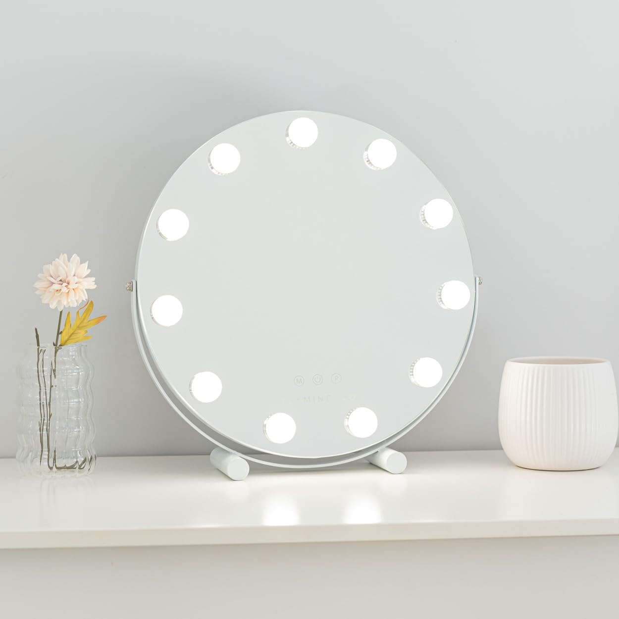 Un espejo de maquillaje blanco redondo con luces, un jarrón con flores y una maceta blanca están sobre una superficie blanca contra una pared gris.