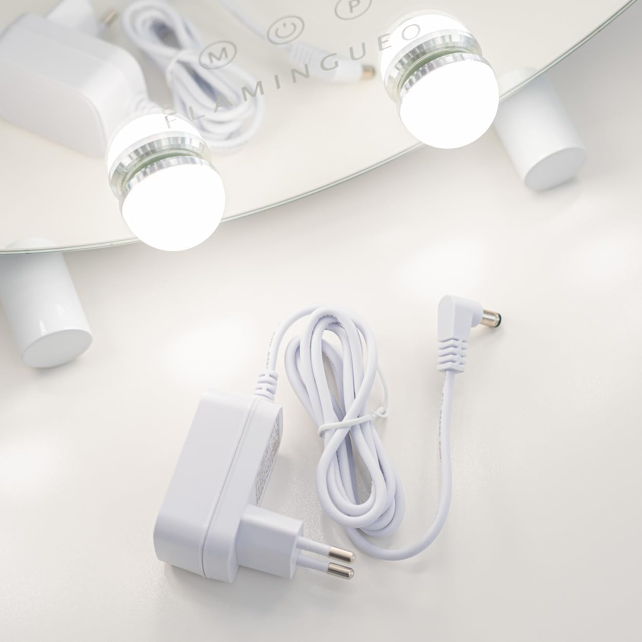 Espejo de maquillaje blanco con luces, adaptadores de corriente y cables sobre una superficie blanca. El espejo está encendido.