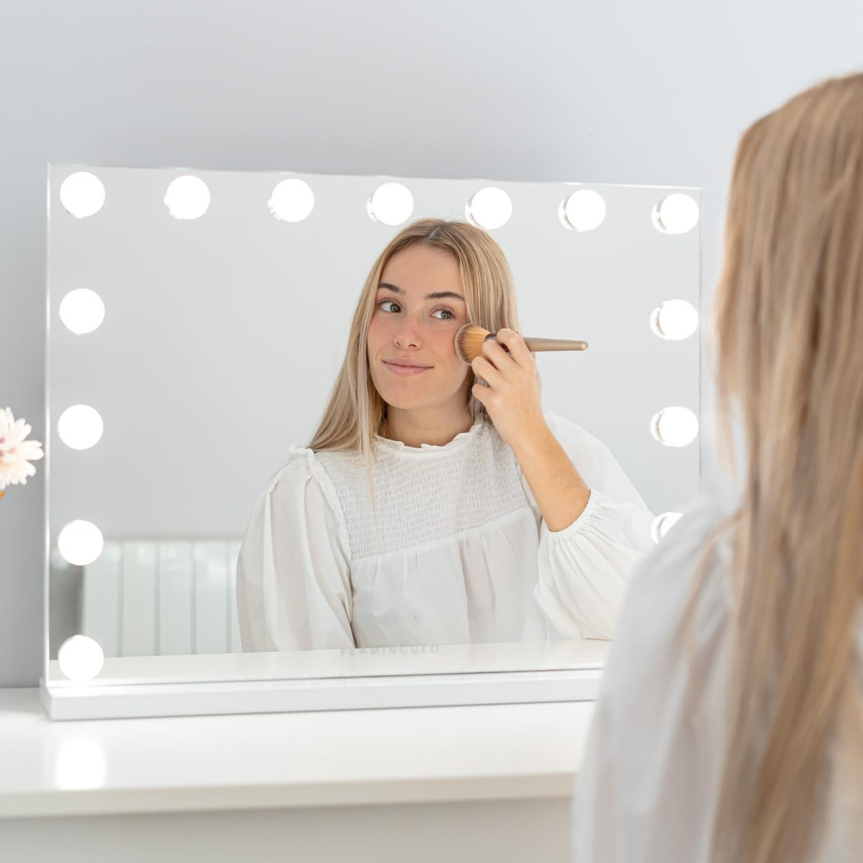 Una mujer se maquilla frente a un espejo. El espejo tiene luces alrededor. Lleva una camisa blanca. Su reflejo es visible.
