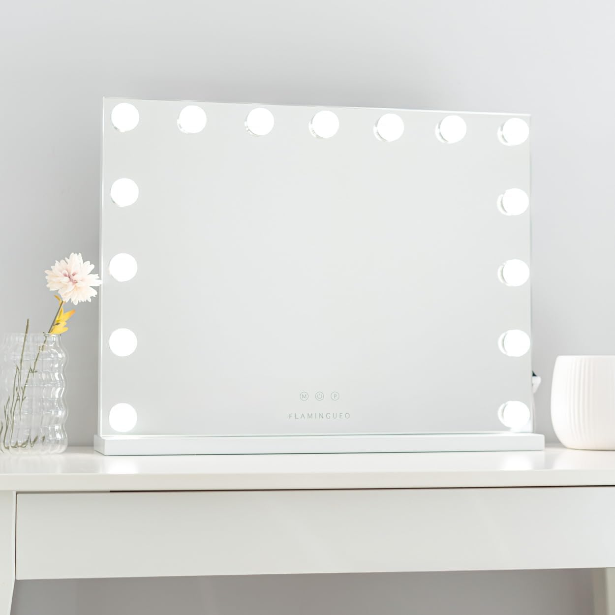 Un espejo de tocador blanco con luces alrededor del borde está sobre una mesa blanca, junto a una flor en un florero y una taza blanca.