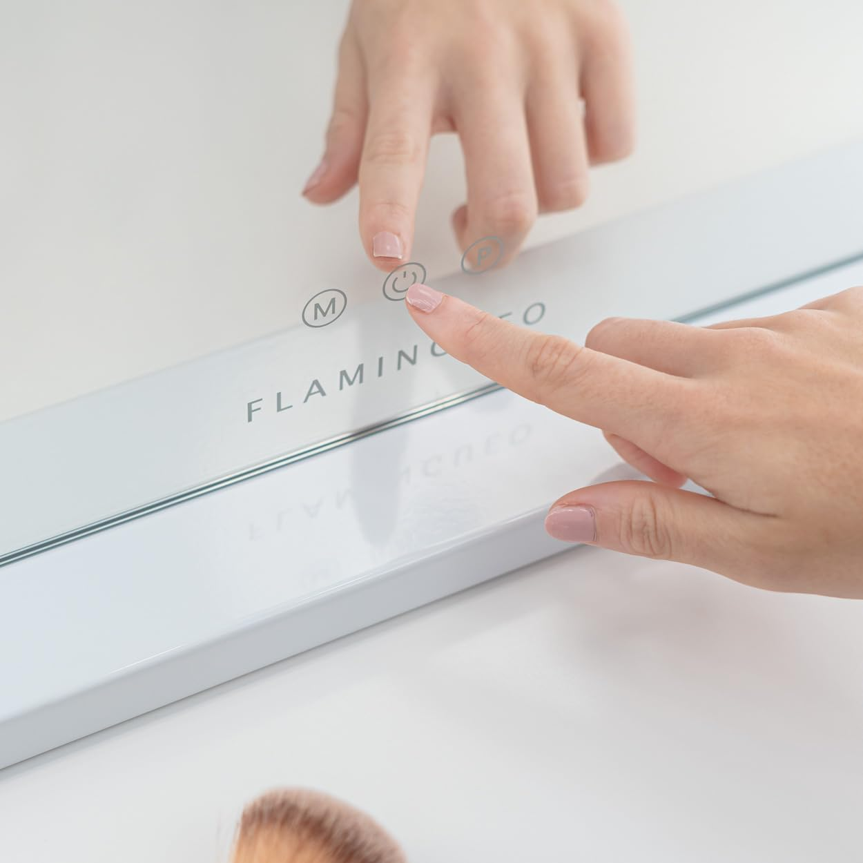 Una mano presiona el botón de encendido en un espejo blanco. El espejo dice 'FLAMINGO'. Hay un pincel de maquillaje abajo.