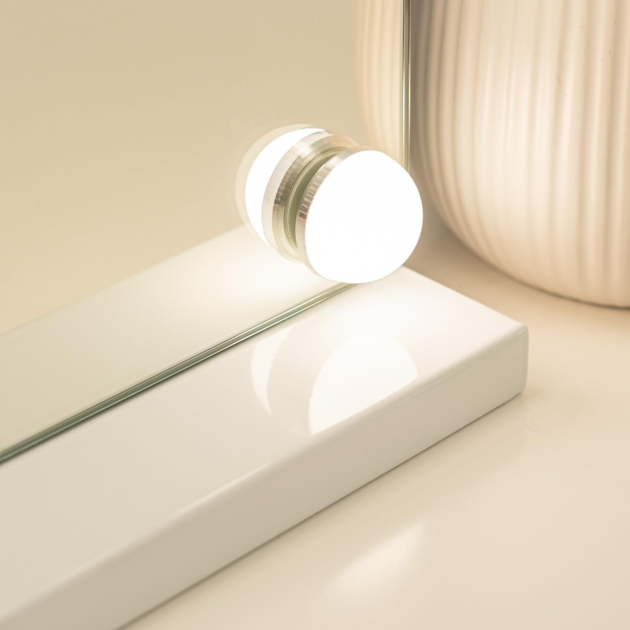 Primer plano de un espejo de maquillaje iluminado con base blanca. Una jarra blanca en el fondo. La escena está iluminada con luz cálida.