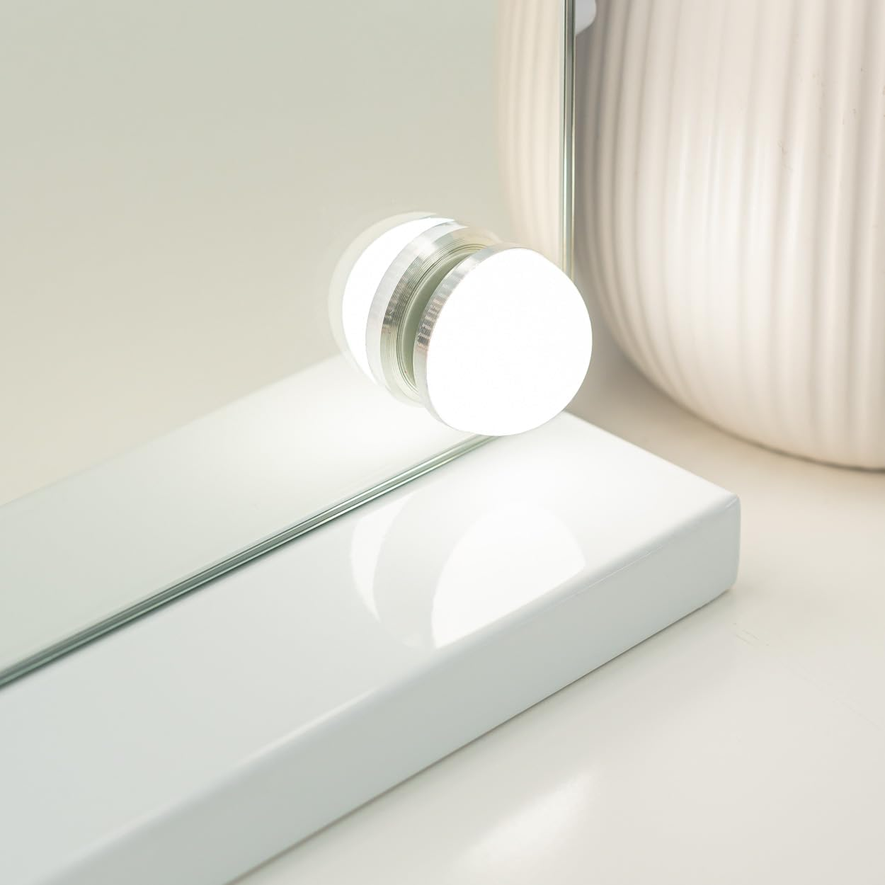 Primer plano de un espejo de maquillaje blanco con luz redonda, una mesa blanca y un jarrón blanco acanalado.