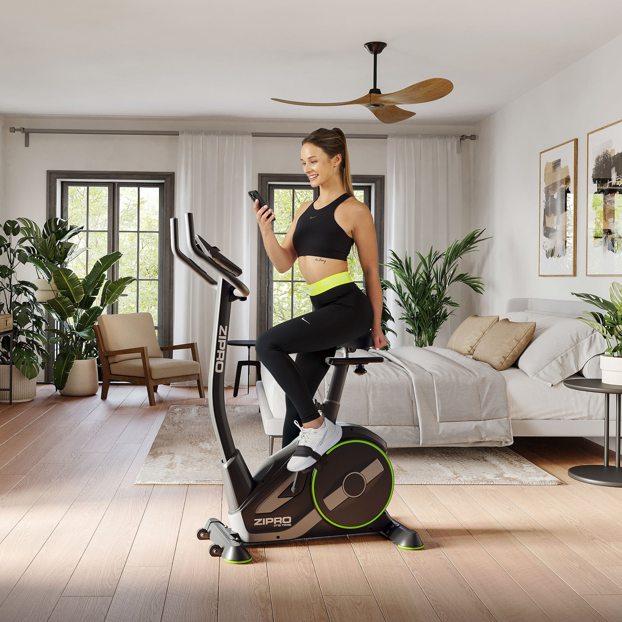 Mujer en bicicleta estática ZIPRO, usando teléfono en habitación luminosa. Cama, plantas y arte al fondo.