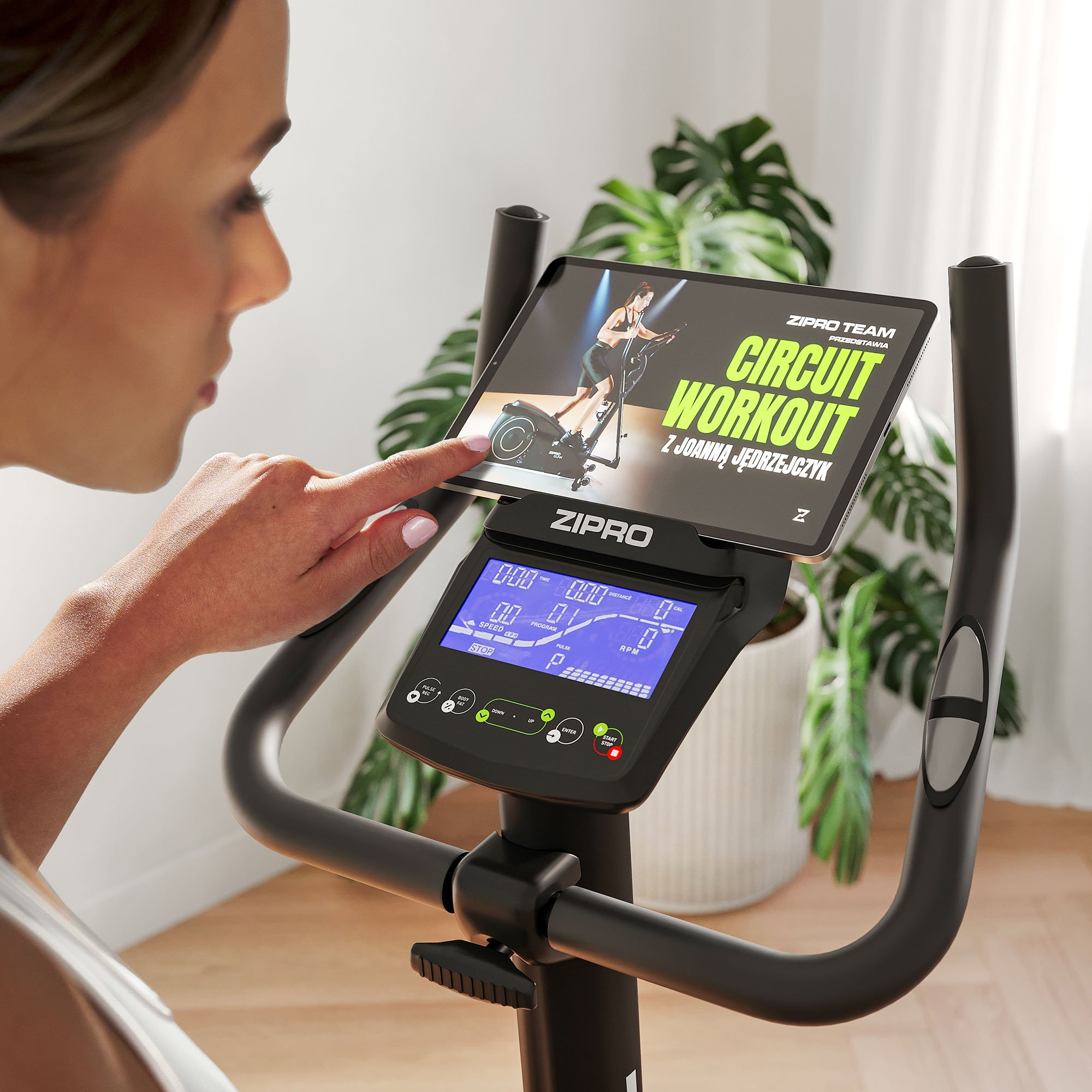 Mujer usa una tableta en una bicicleta estática. La pantalla de la tableta muestra un video de entrenamiento. La pantalla muestra métricas. Plantas en el fondo.