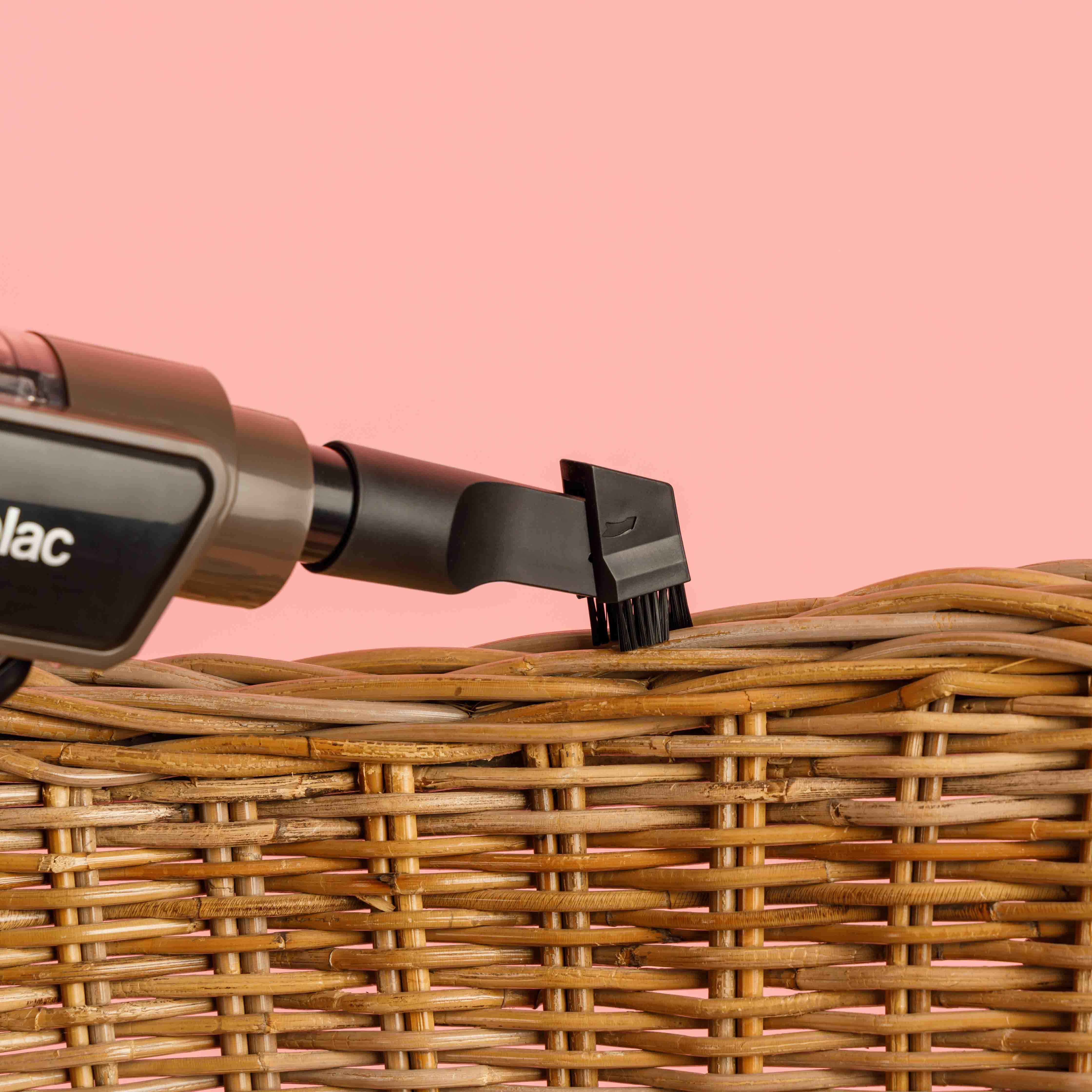 Odkurzacz ręczny czyści wiklinowy kosz na różowym tle. Odkurzacz ma końcówkę szczotkową.