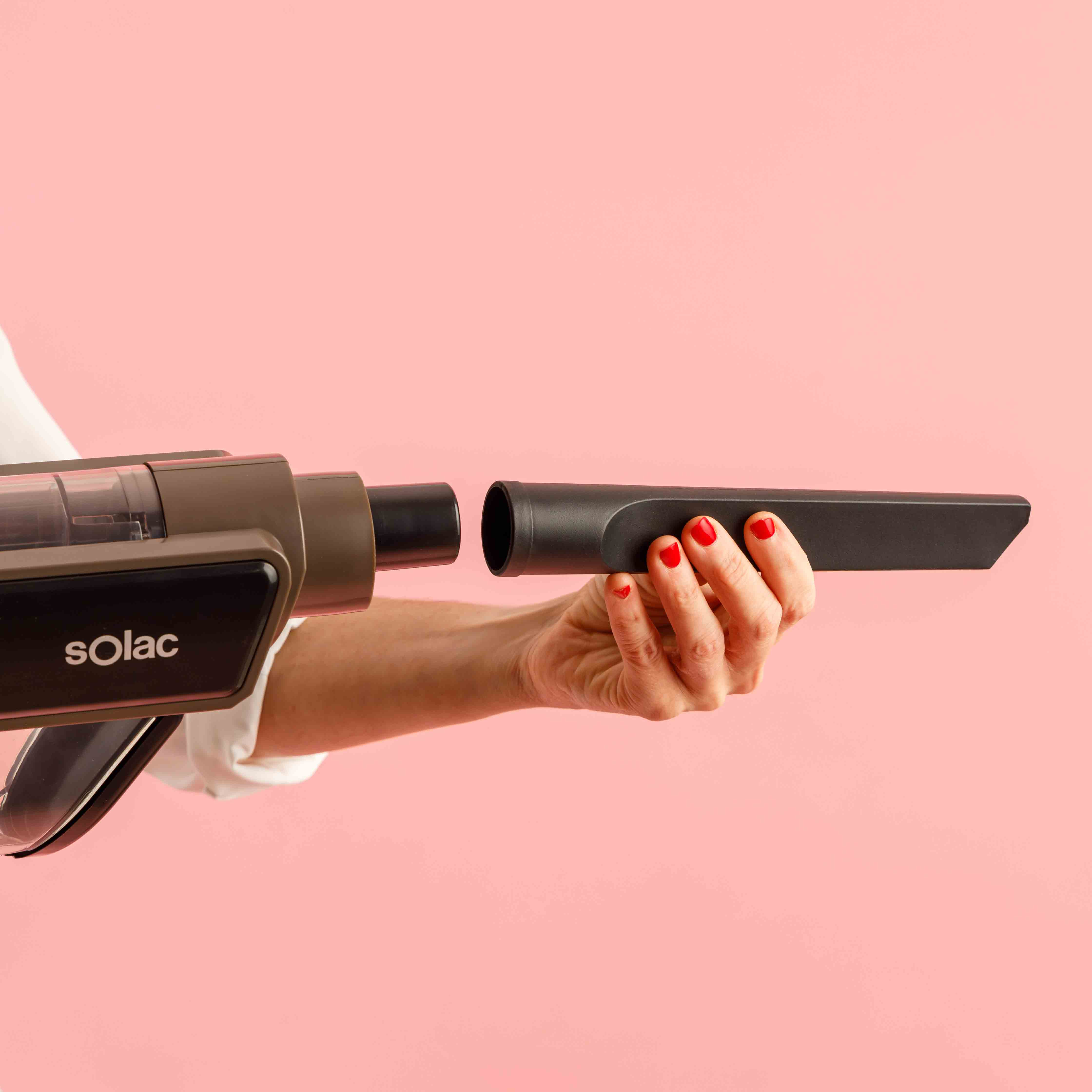 Ręka trzyma czarną nasadkę do odkurzacza w pobliżu odkurzacza. Tło jest różowe.