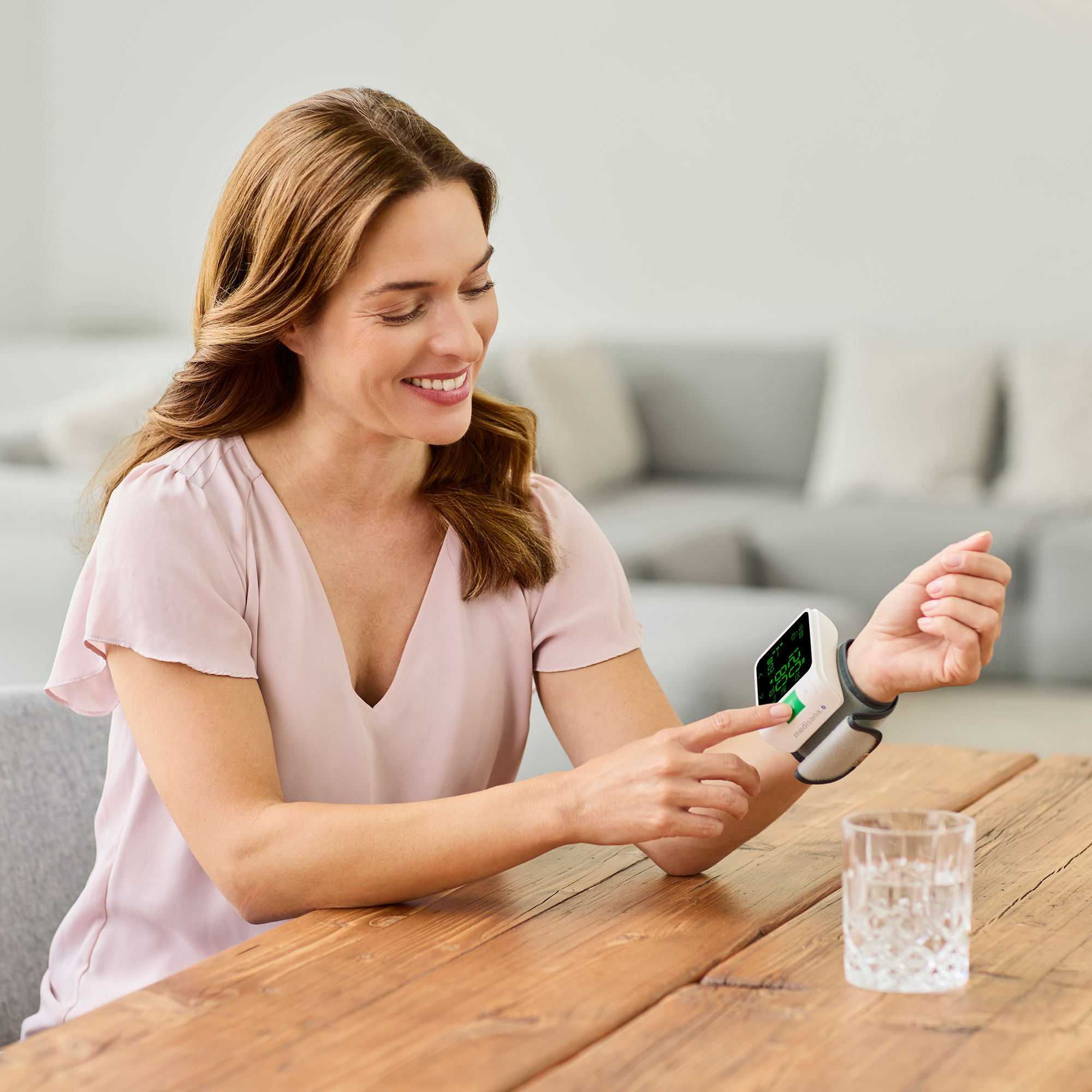 Frau misst Blutdruck mit Monitor am Handgelenk. Ein Glas Wasser steht auf dem Holztisch.