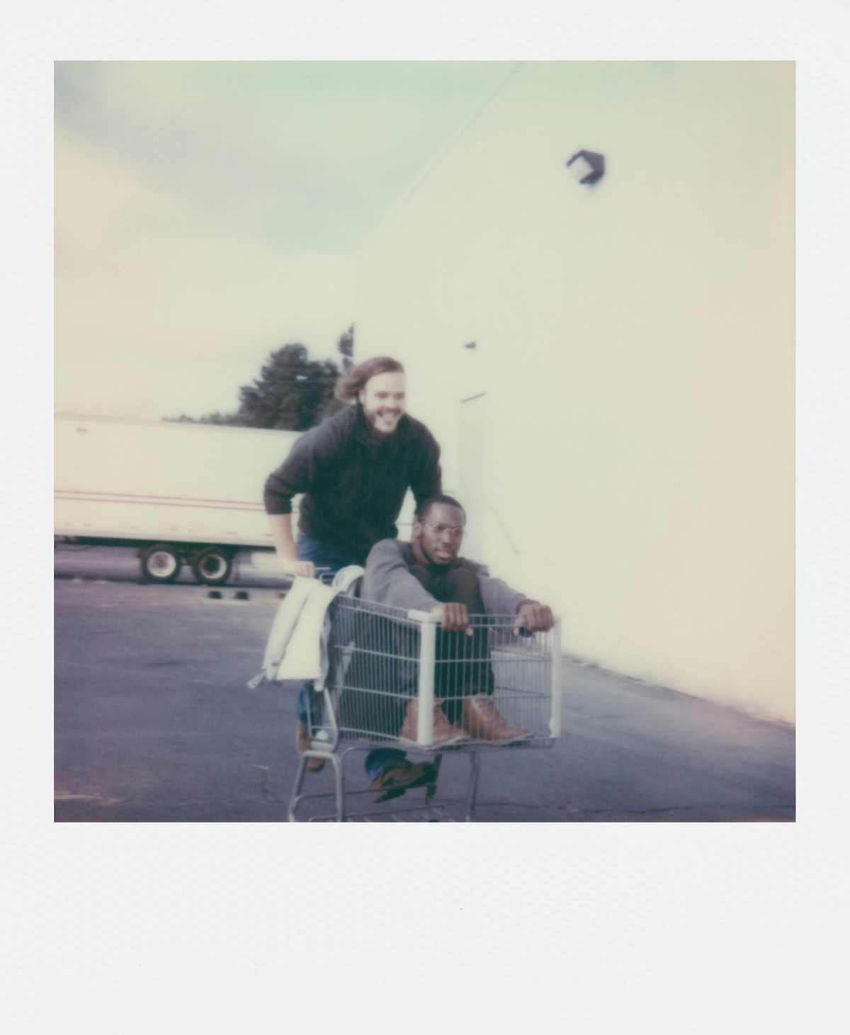 Dwóch mężczyzn, jeden w wózku sklepowym, drugi pcha. Są na zewnątrz, w pobliżu białego budynku i ciężarówki.