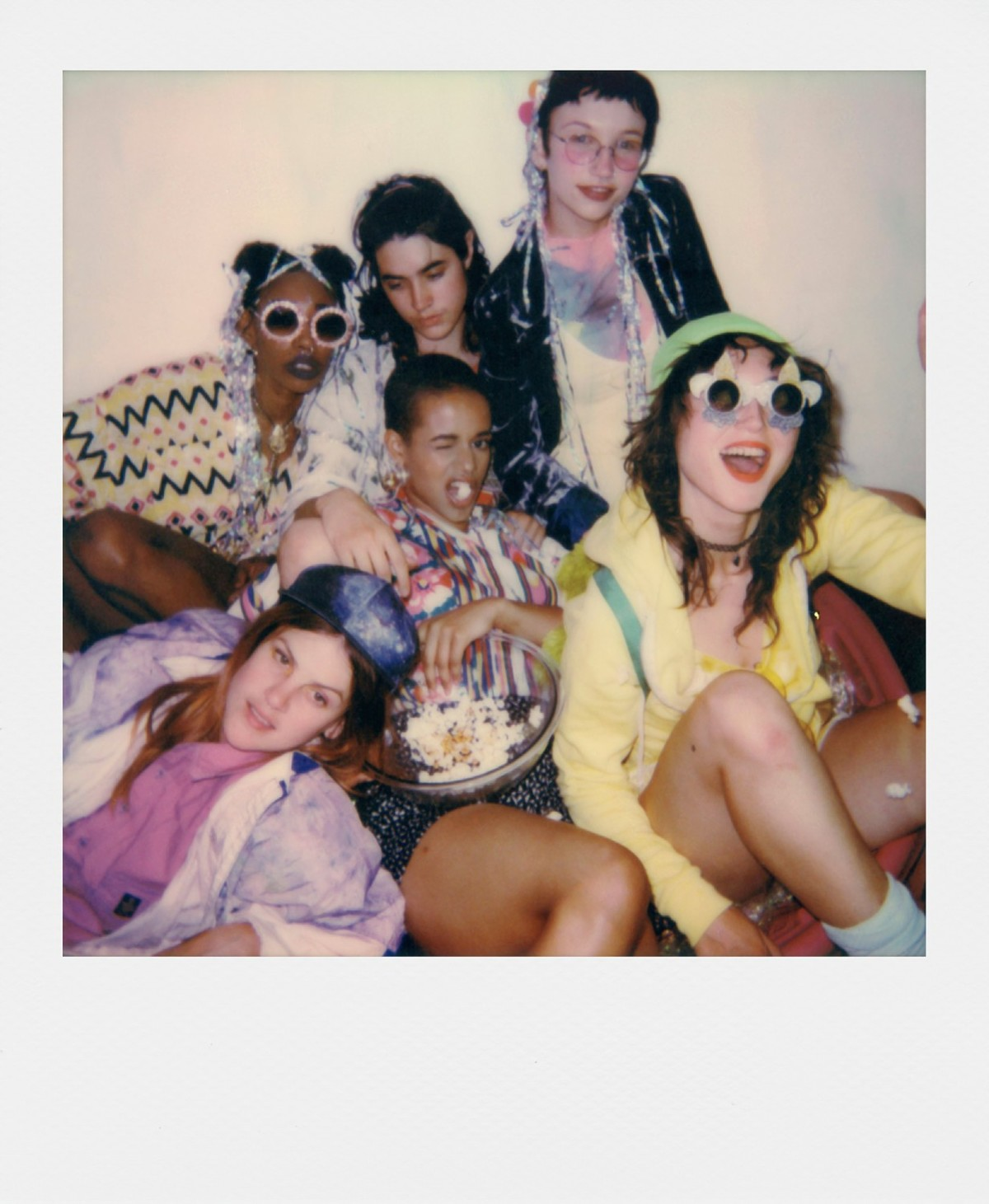 Grupa młodych ludzi w kolorowych strojach i okularach przeciwsłonecznych, niektórzy jedzą popcorn, pozują i uśmiechają się na kanapie.