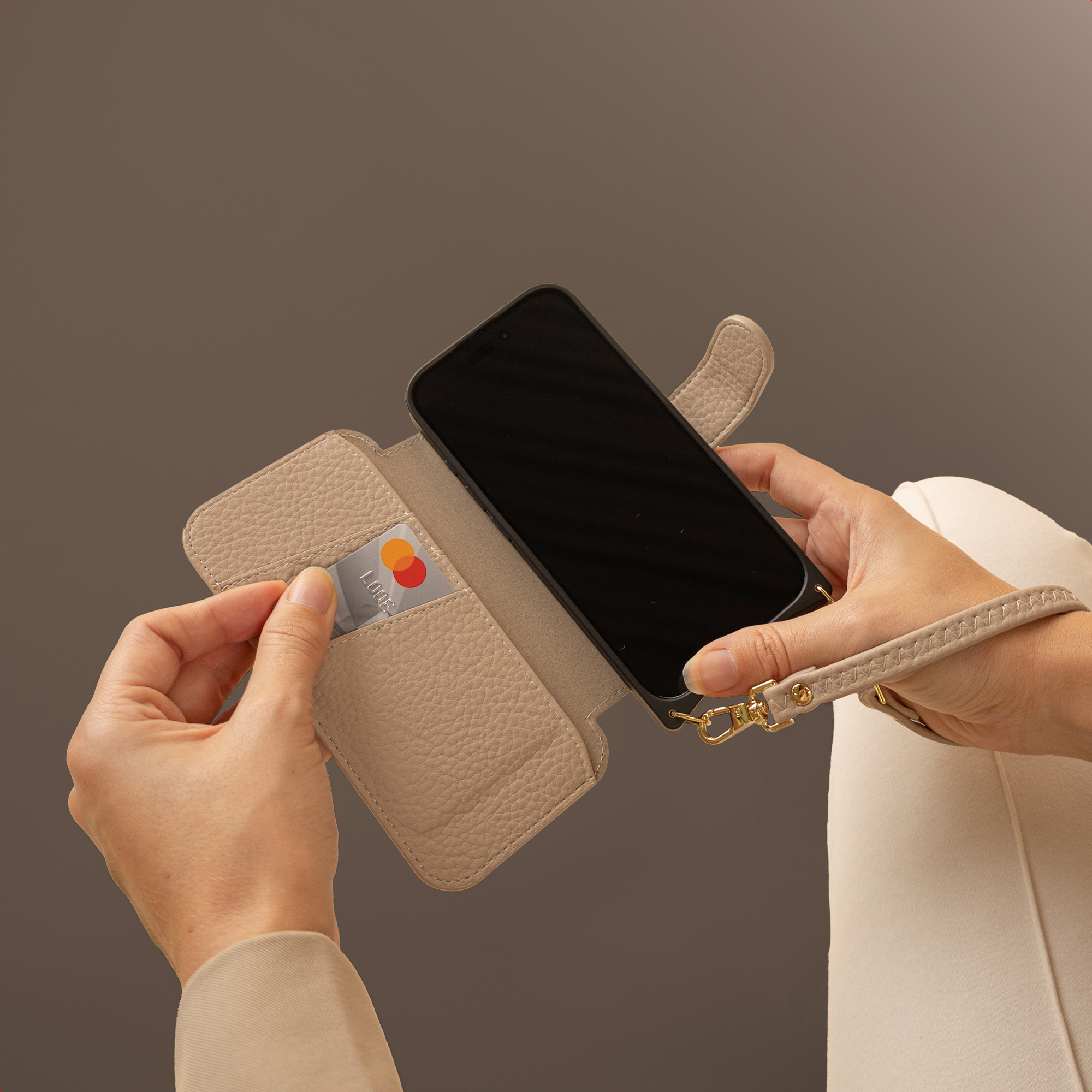 Mains tenant un téléphone dans un étui en cuir beige. Une carte de crédit est placée dans une poche.