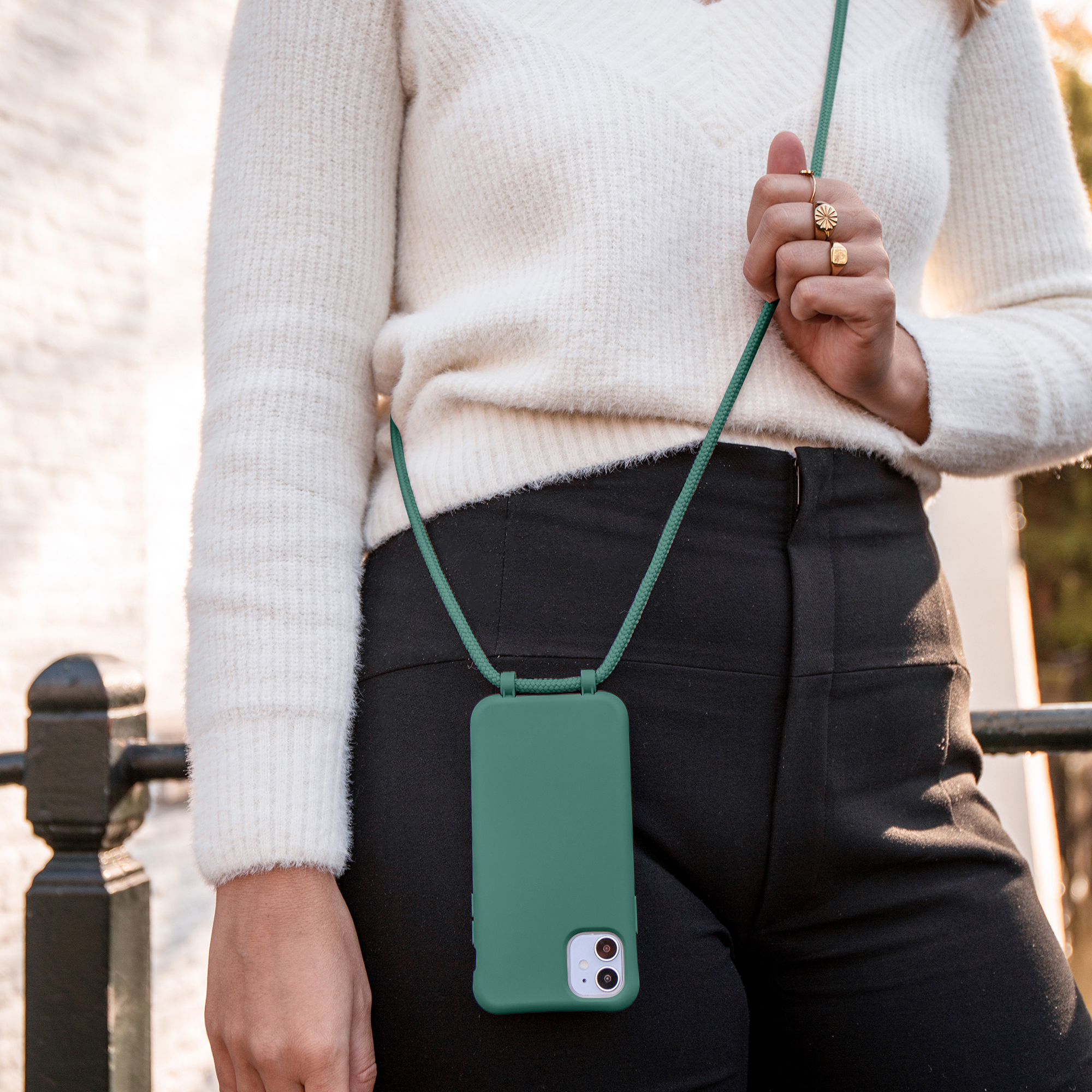Een vrouw draagt een witte trui en een zwarte broek en houdt een groene telefoonhoes met bijpassend koord vast. Ze heeft ringen om haar vingers.
