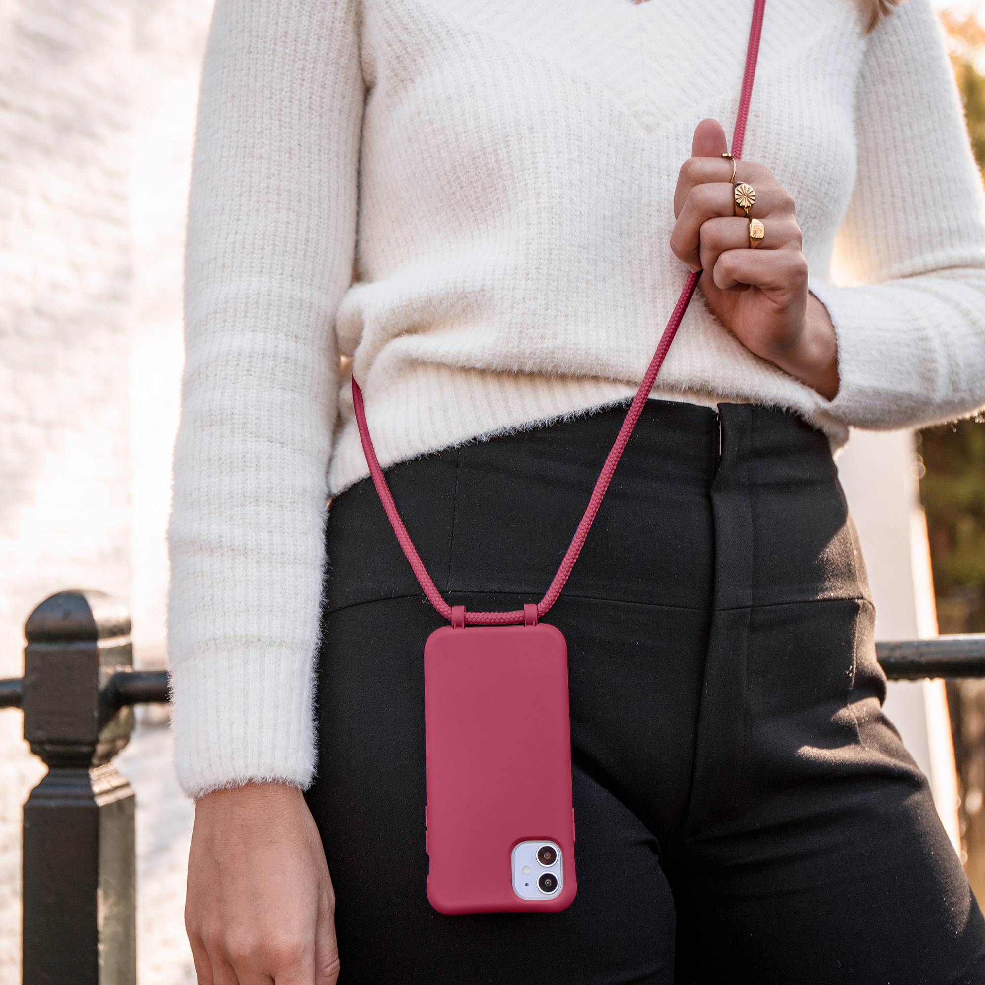 Een persoon draagt een witte trui en een zwarte broek met een rode telefoonhoes. De hoes hangt aan een rood koord.