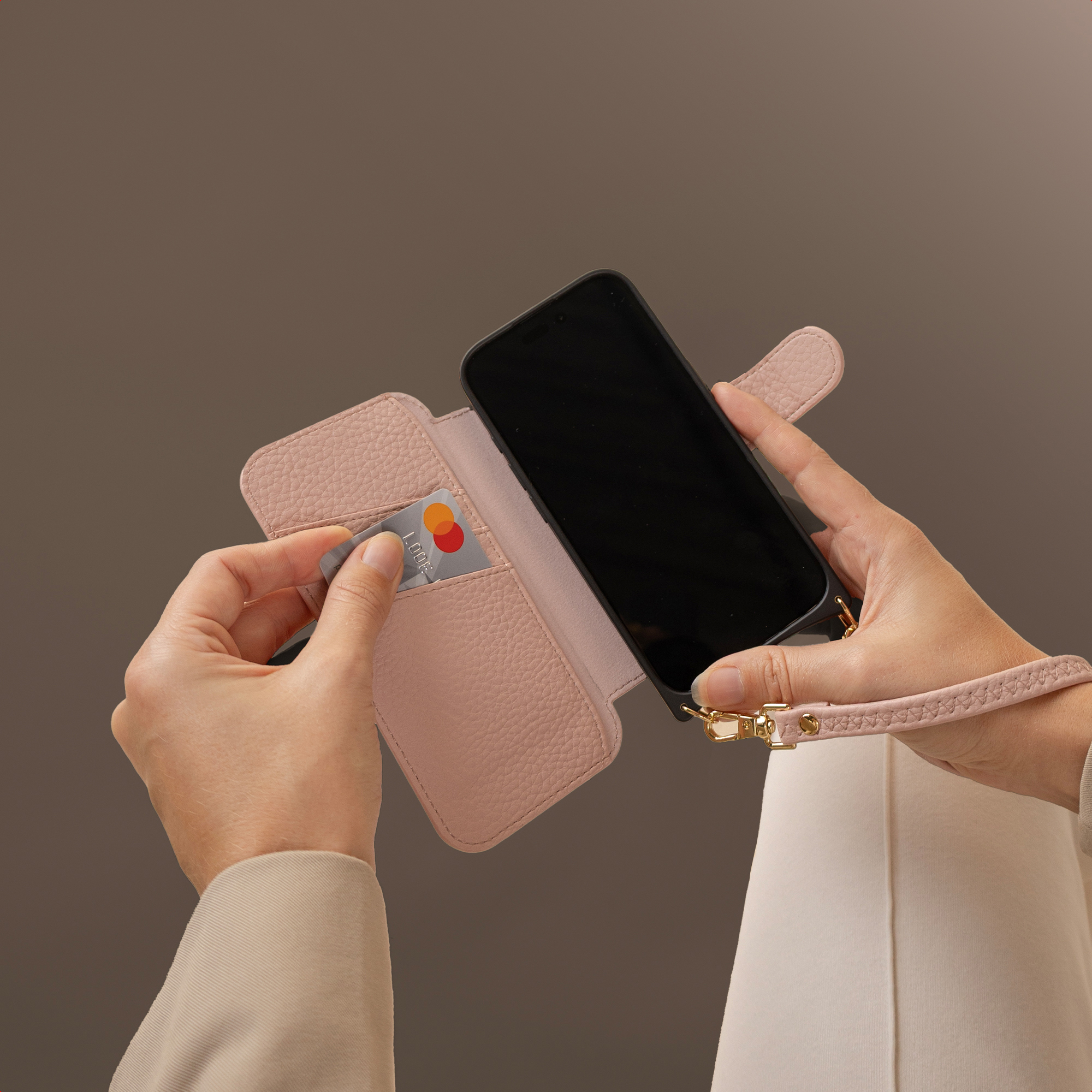 Een persoon houdt een roze telefoonhoes vast met een creditcard erin. Er zit een zwarte telefoon in de hoes. De achtergrond is bruin.