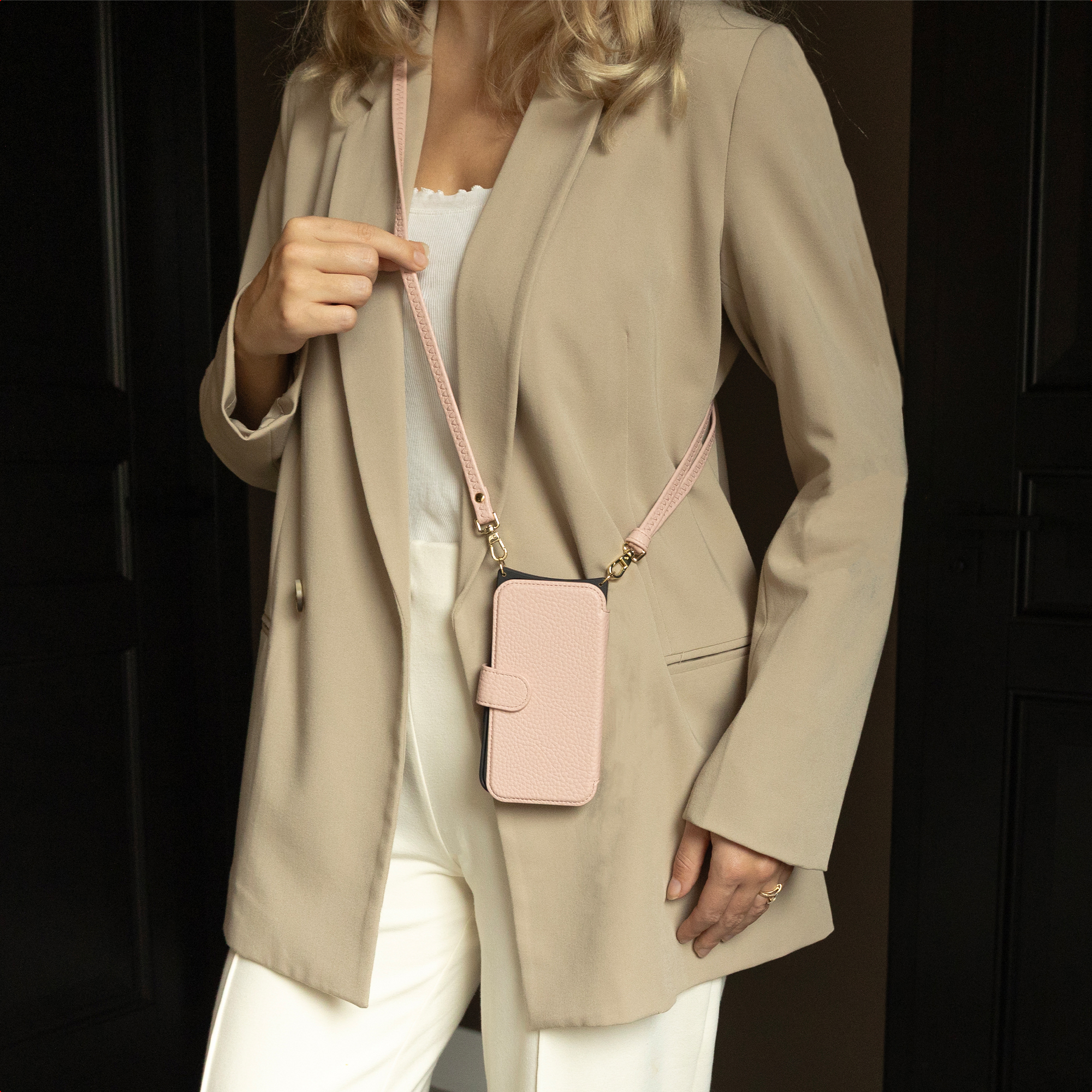 Een vrouw in een beige blazer en witte broek houdt een roze telefoonhoesje met een band vast. Op de achtergrond een donkere houten deur.