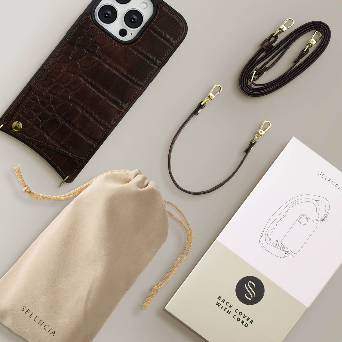 Een bruine leren telefoonhoes, tas en koord worden getoond. Een witte doos en een zakje met trekkoord zijn ook te zien, allemaal op een grijs oppervlak.