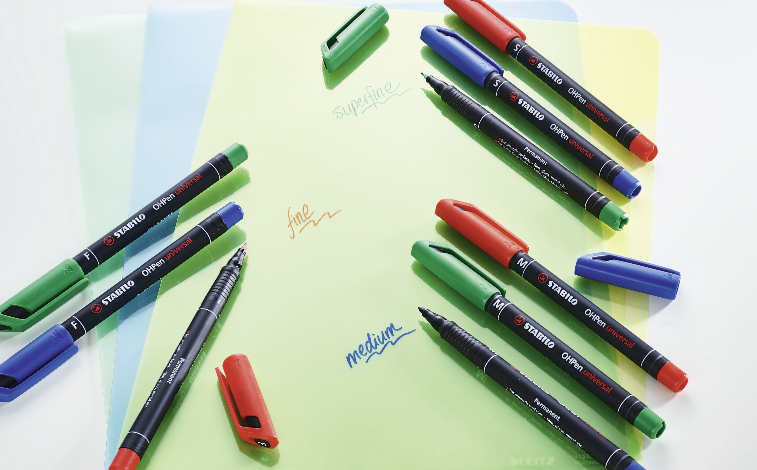 Das Bild zeigt mehrere STABILO OHPen Universalmarker in verschiedenen Farben mit geöffneten Kappen. Der Hintergrund ist hellgrün und blau.