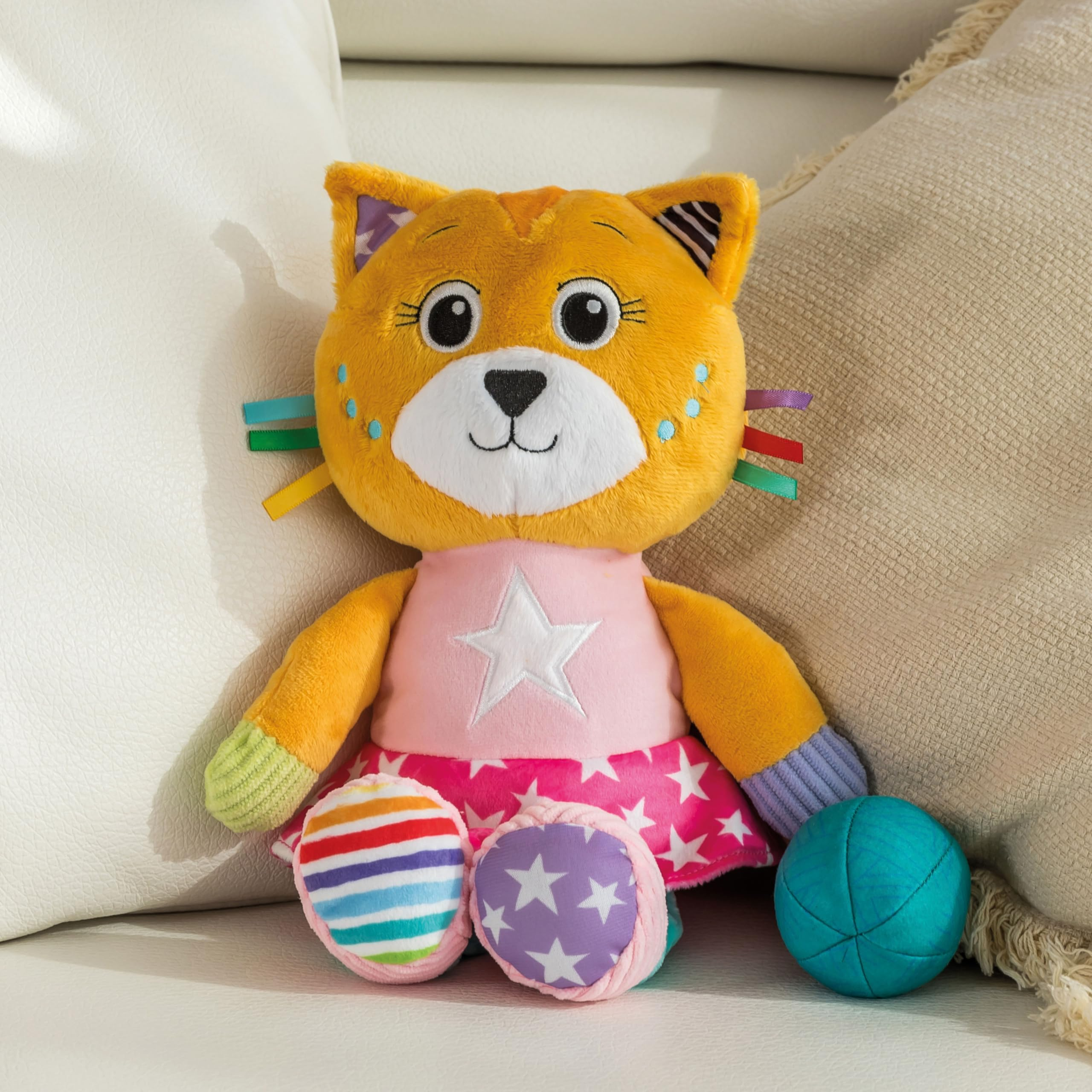 Peluche di gatto arancione con vestito rosa e stella bianca, su superficie bianca con palla blu. Cuscino beige sullo sfondo.