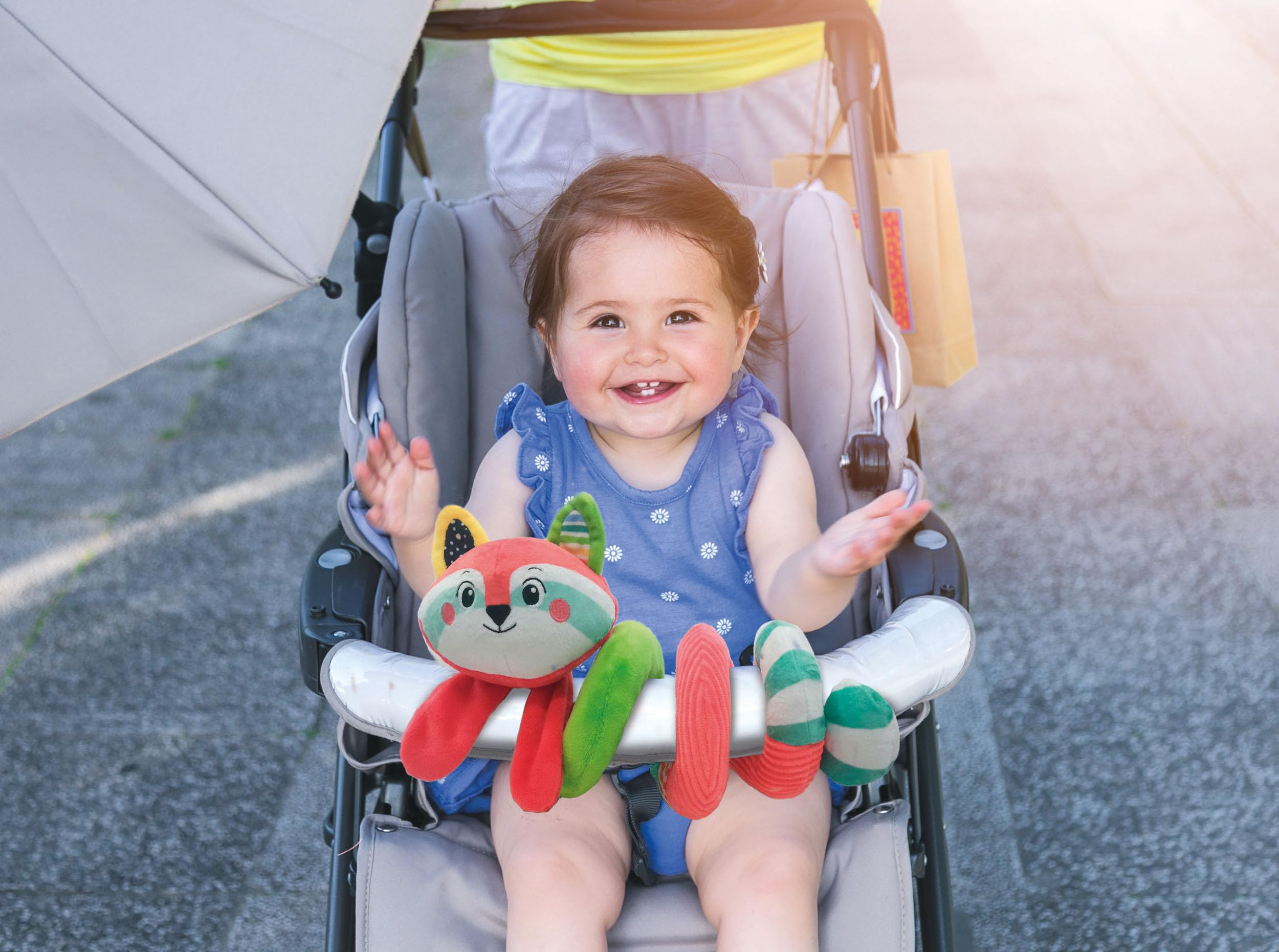 Un bambino in un passeggino sorride, tenendo un giocattolo. Un ombrello è aperto. Una persona è dietro.