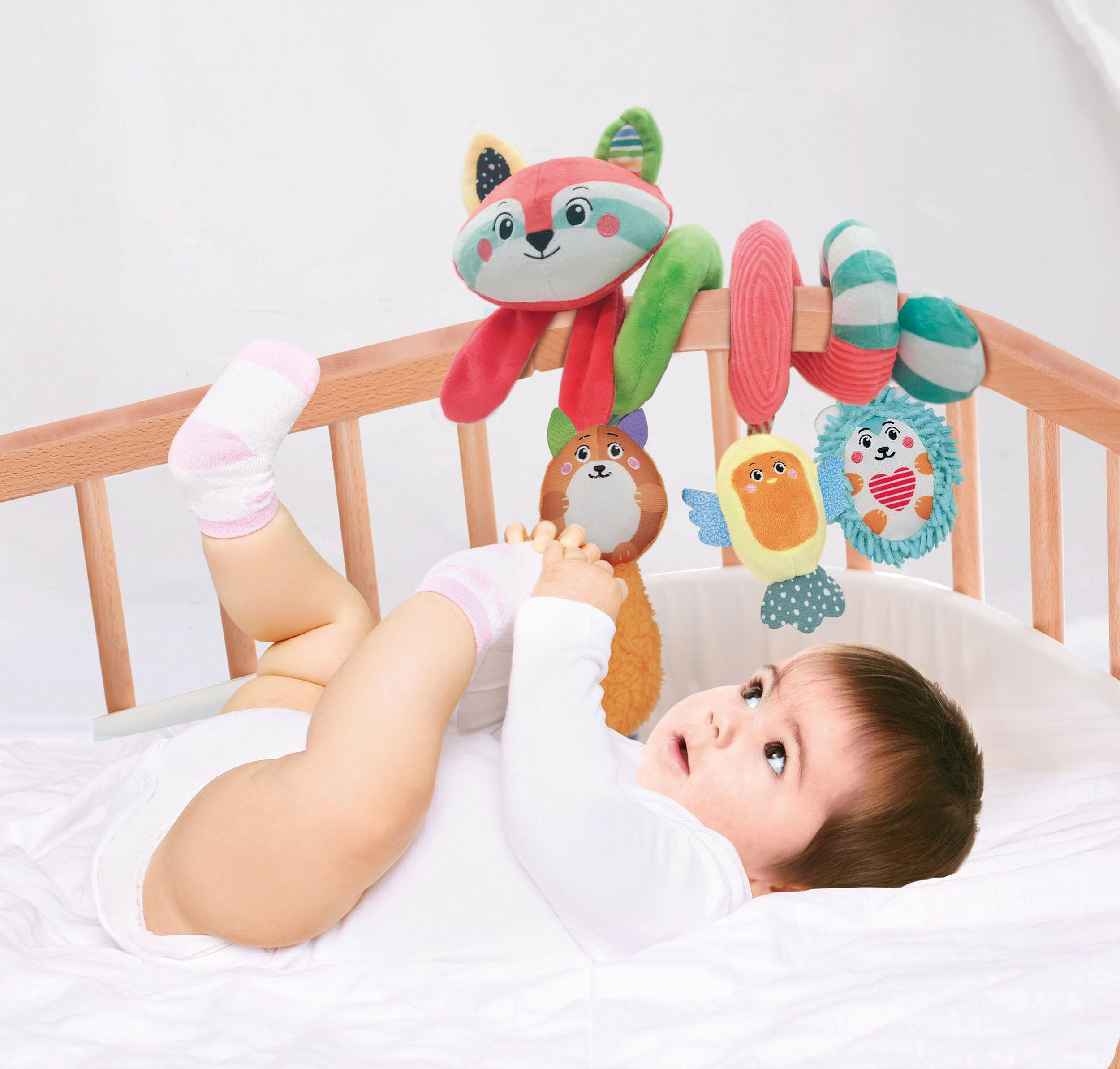 Un bambino in una culla guarda una colorata volpe giocattolo. Il bambino indossa un body bianco e calzini rosa.