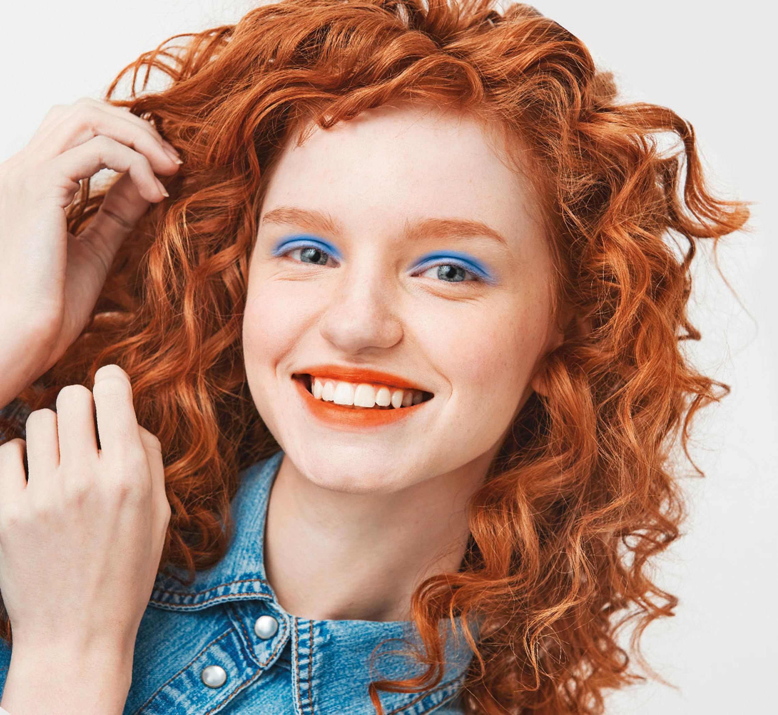 Eine Frau mit roten Locken lächelt. Sie trägt blauen Lidschatten und einen orangen Lippenstift. Sie trägt eine Jeansjacke.