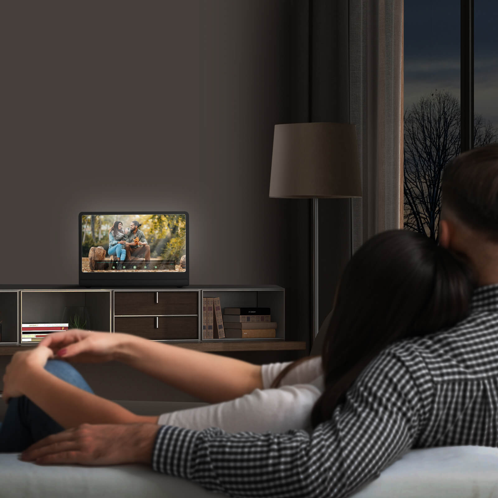 Pareja viendo la televisión. TV muestra una pareja. Lámpara y ventana al fondo. Habitación oscura.