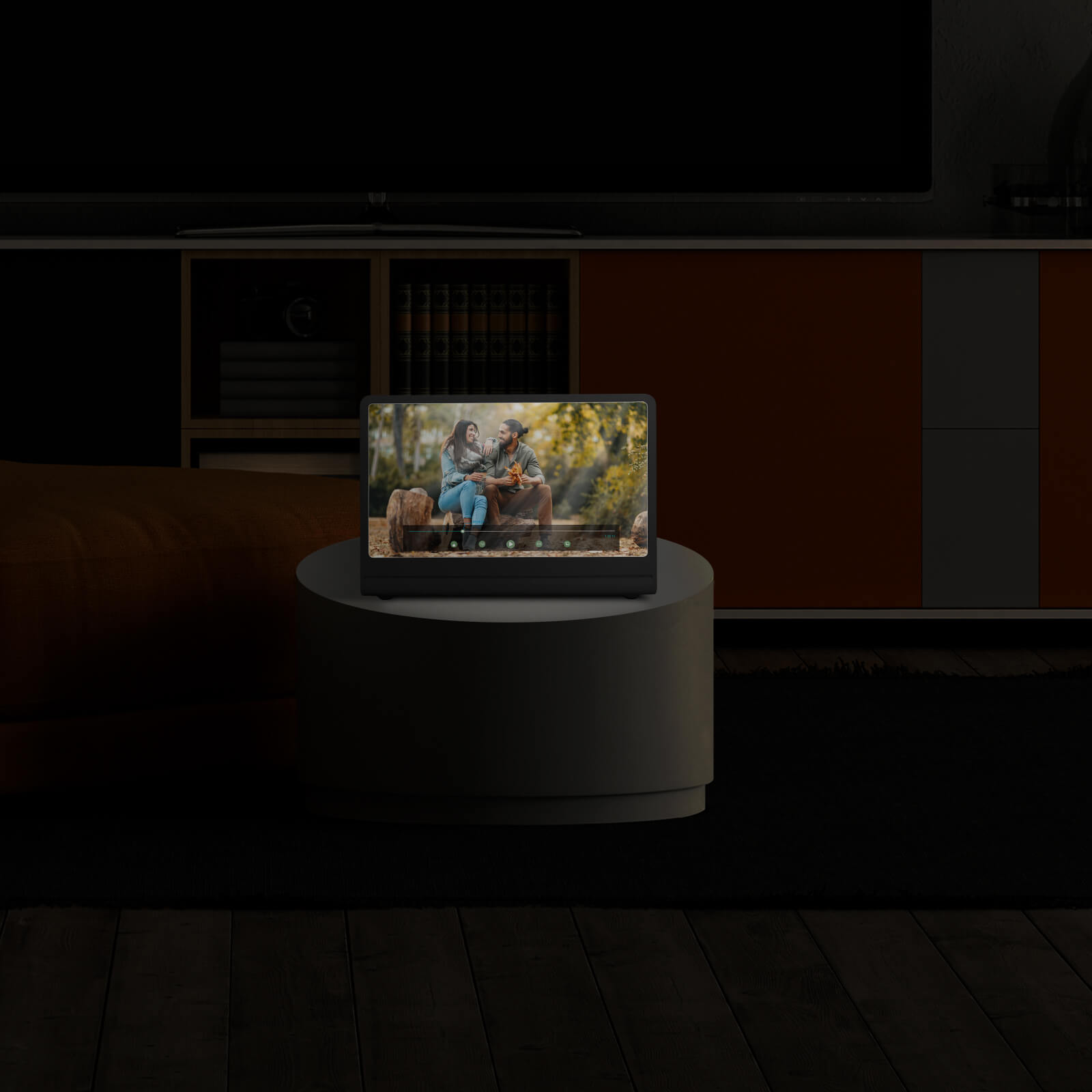 Una tableta muestra una pareja al aire libre. Está sobre una mesa redonda, frente a un armario y un televisor. Habitación oscura.