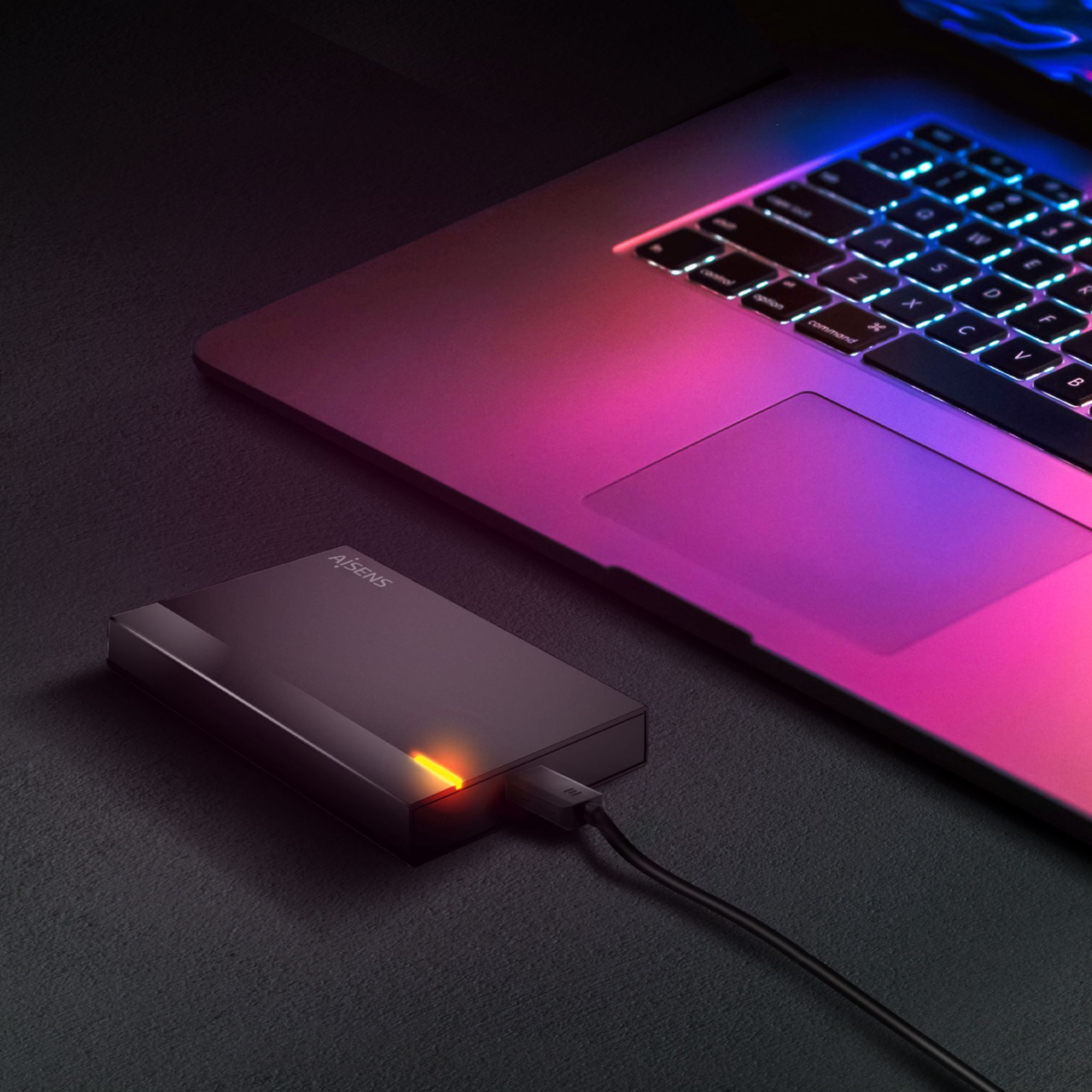 Una laptop y un disco duro externo sobre una superficie oscura. El teclado de la laptop está iluminado. El disco está conectado.