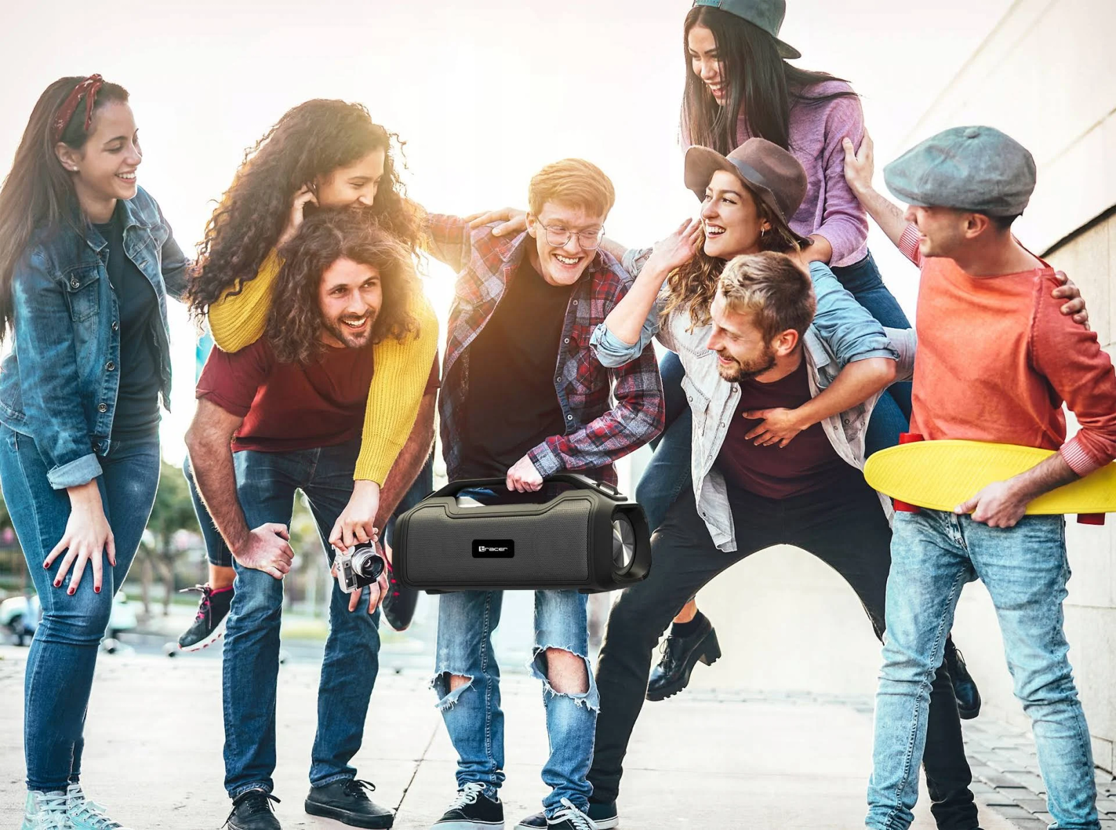 Gruppo di amici che ridono, alcuni a cavalluccio. Uno tiene un altoparlante. Altri hanno uno skateboard. Ambiente esterno luminoso.