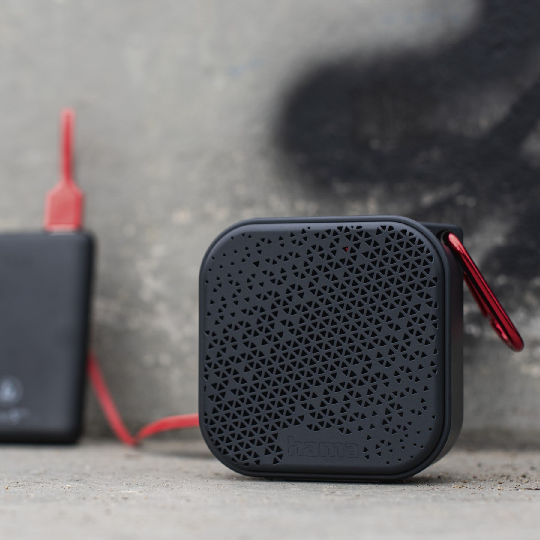 Un altoparlante nero con moschettone e cavo rossi, collegato a un power bank nero, contro un muro grigio.