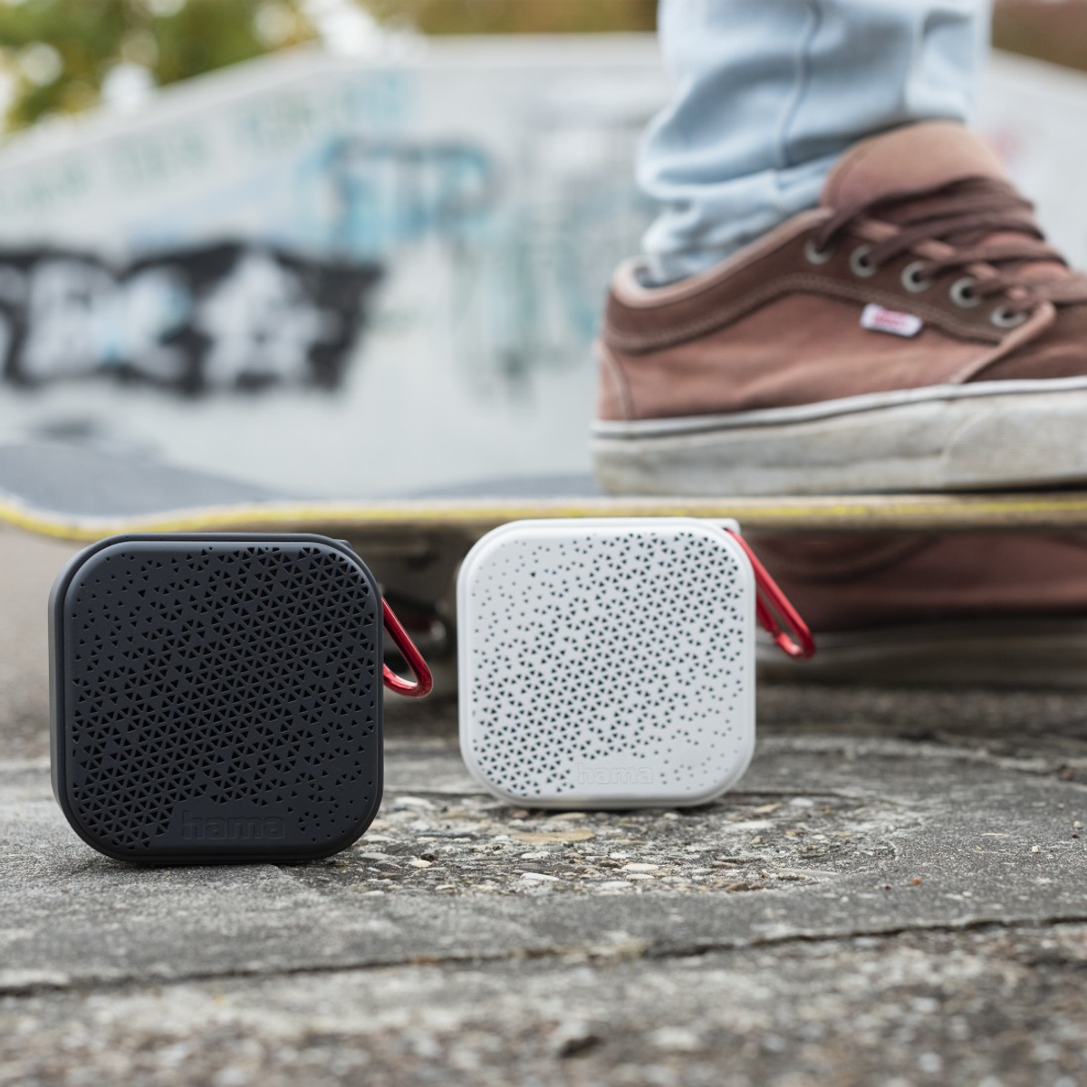 Due altoparlanti quadrati, uno nero e uno bianco, sono su uno skateboard. Una scarpa è sulla tavola. Lo sfondo è un muro di graffiti.