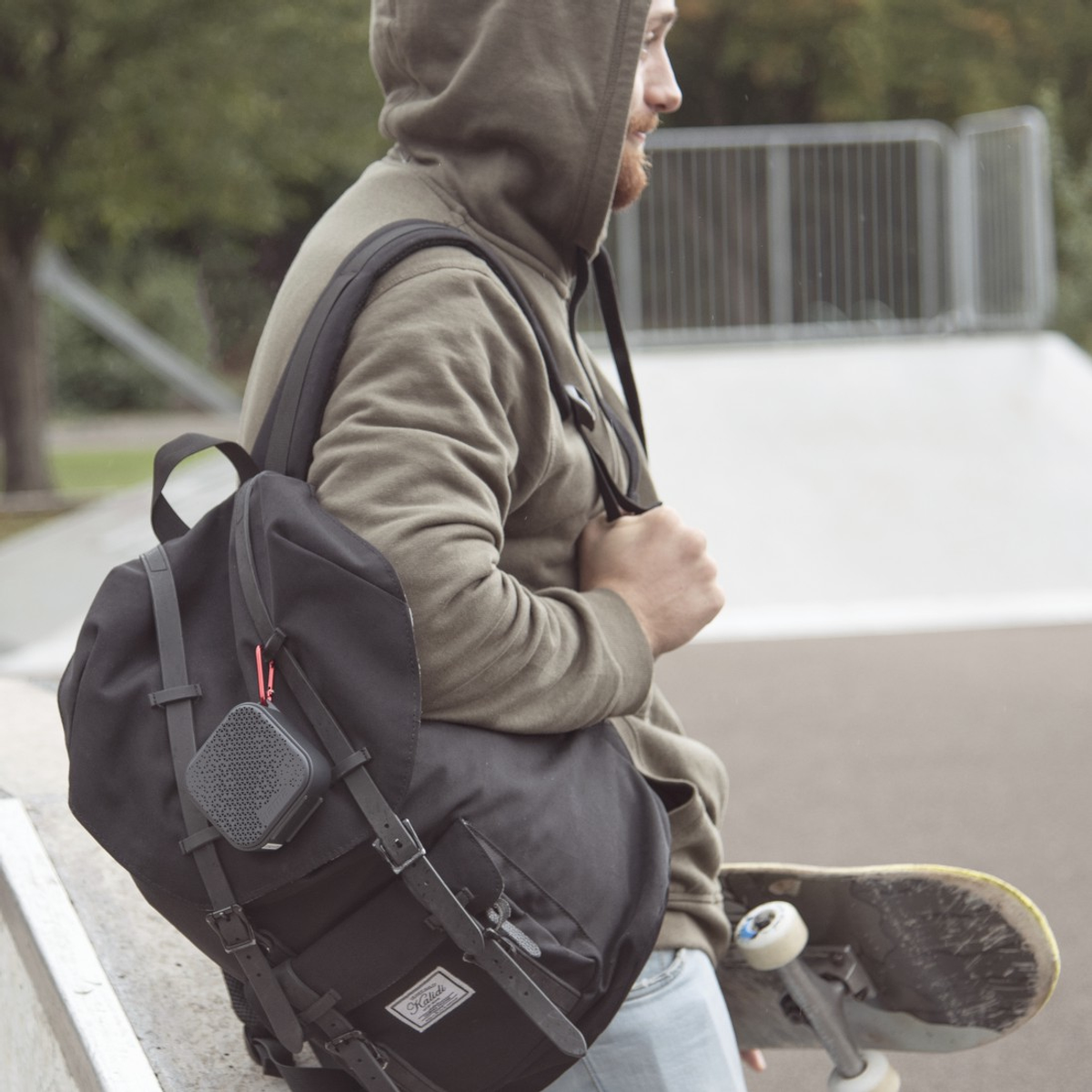 Un uomo con la felpa con cappuccio e uno zaino, tiene uno skateboard. Un altoparlante è appeso alla borsa. È in uno skatepark.