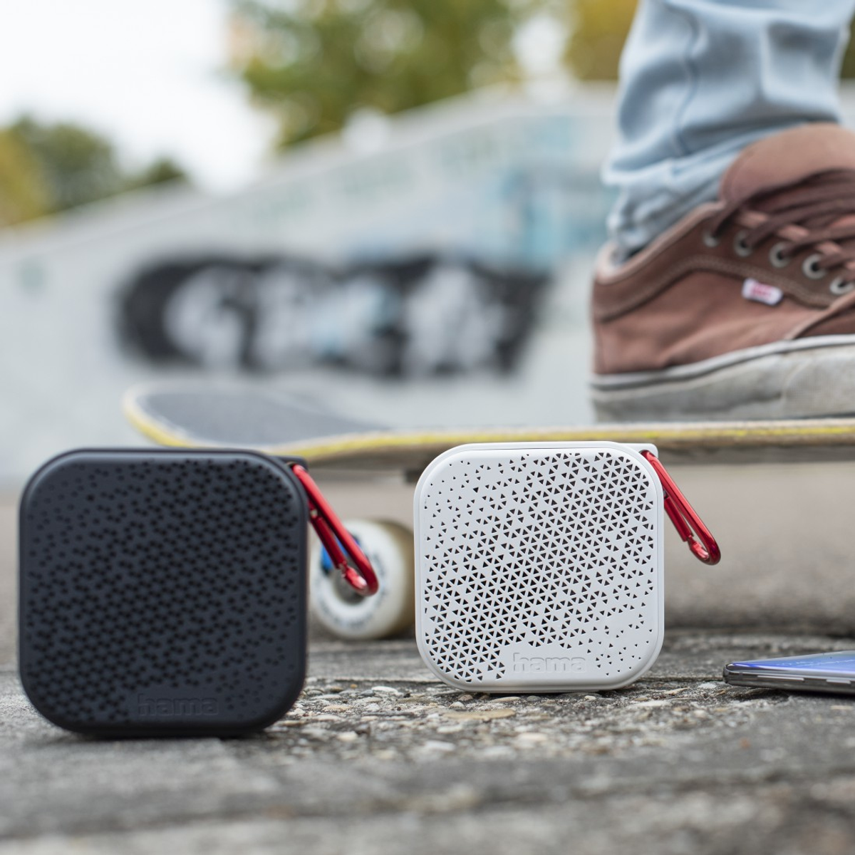 Deux enceintes carrées, une noire, une blanche, avec des clips rouges, sont sur du béton. Un skateboard et une chaussure en arrière-plan.
