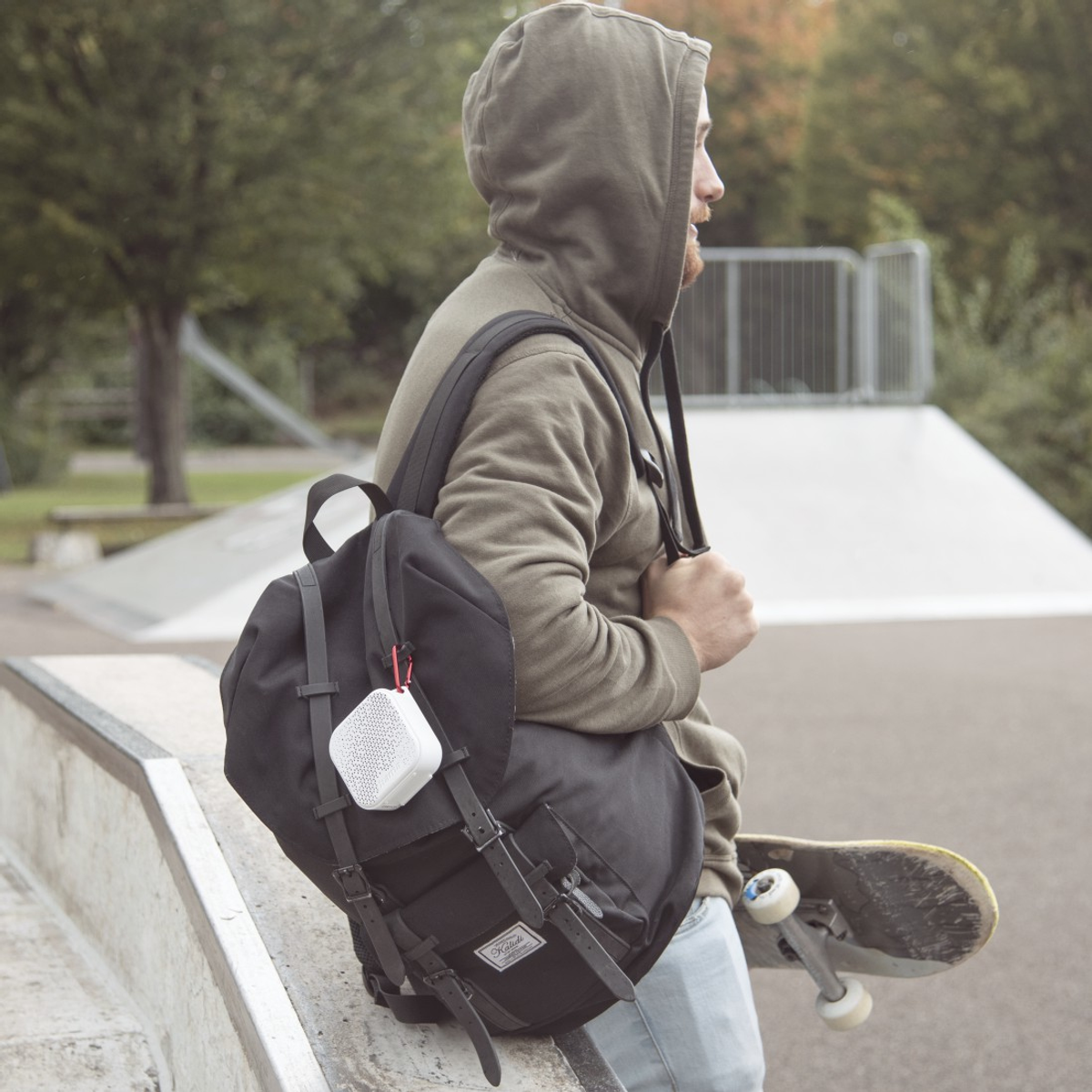Un homme avec un sac à dos et un skateboard dans un skatepark. Il porte un sweat à capuche et un haut-parleur sur le sac.