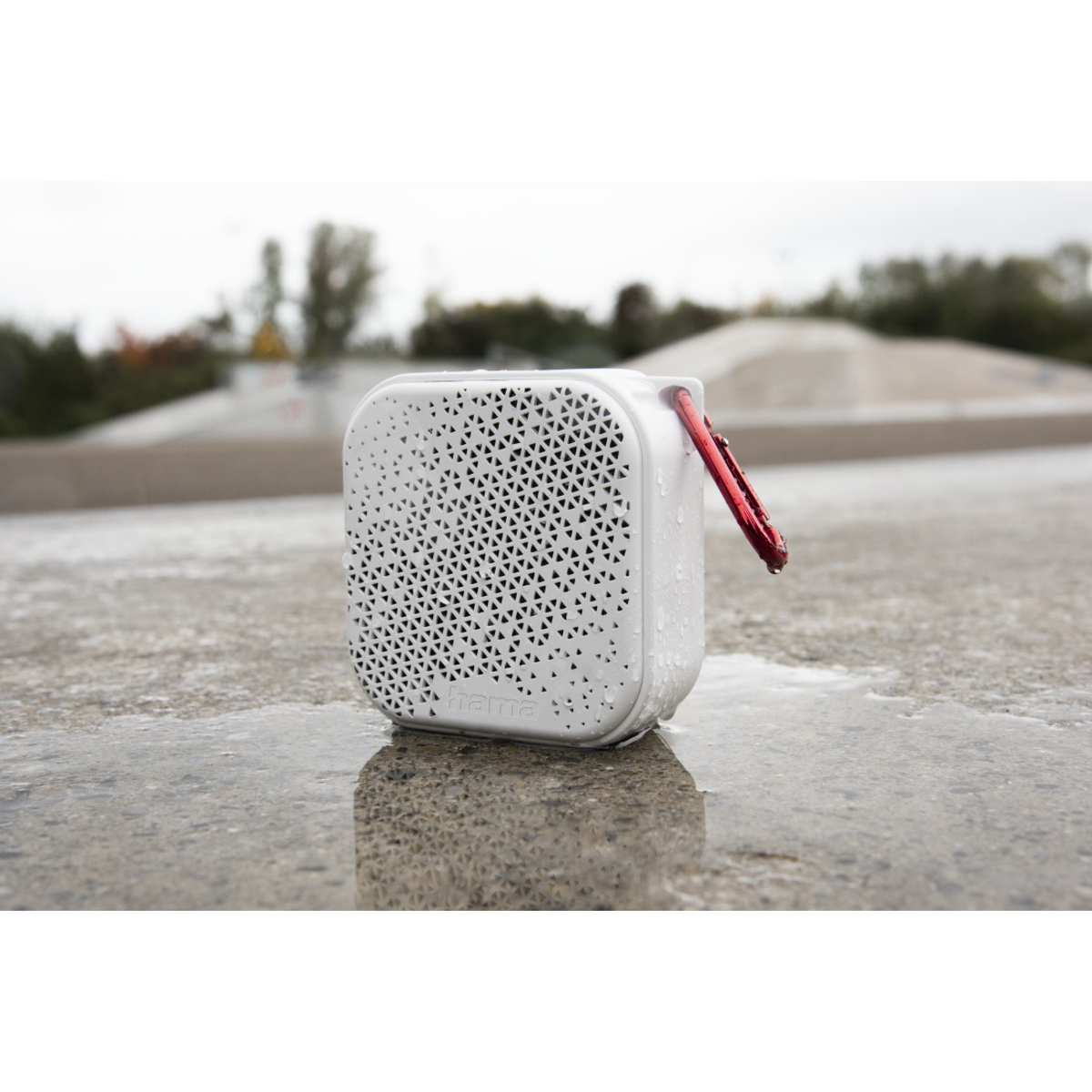 Haut-parleur blanc avec mousqueton rouge sous la pluie. Posé sur du béton mouillé, avec des arbres flous et un skate park en arrière-plan.