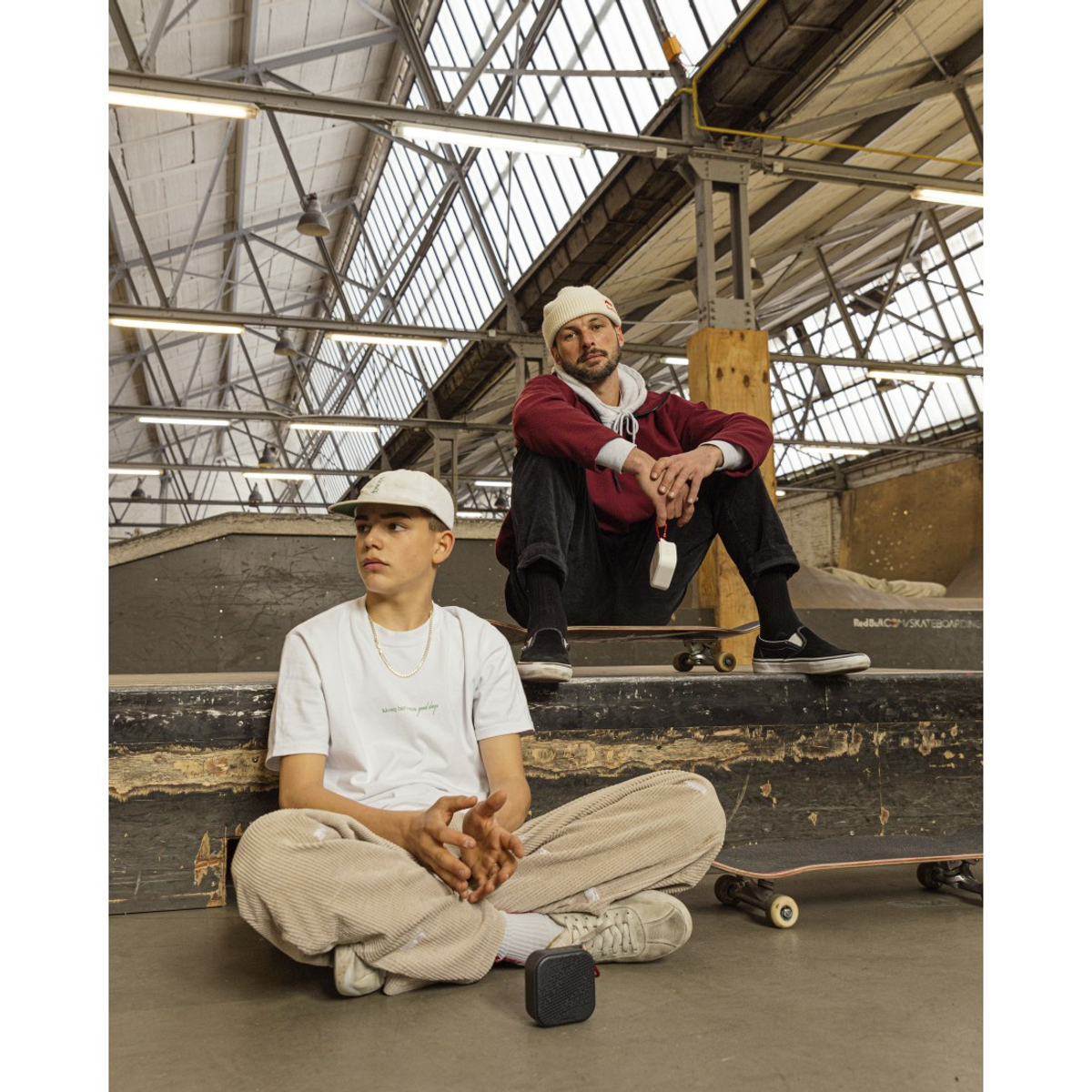 Deux hommes sont assis dans un skatepark. L'un porte une casquette et une chemise blanches. L'autre porte un sweat à capuche rouge. Des skateboards et un haut-parleur sont visibles.