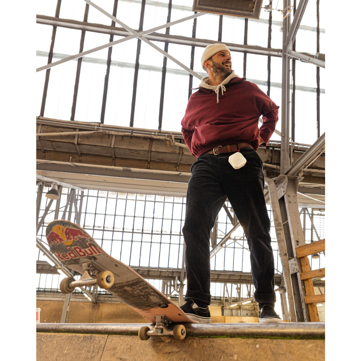 Un homme en sweat à capuche bordeaux et bonnet se tient près d'un skateboard. Il sourit. L'arrière-plan présente des structures métalliques et des fenêtres.