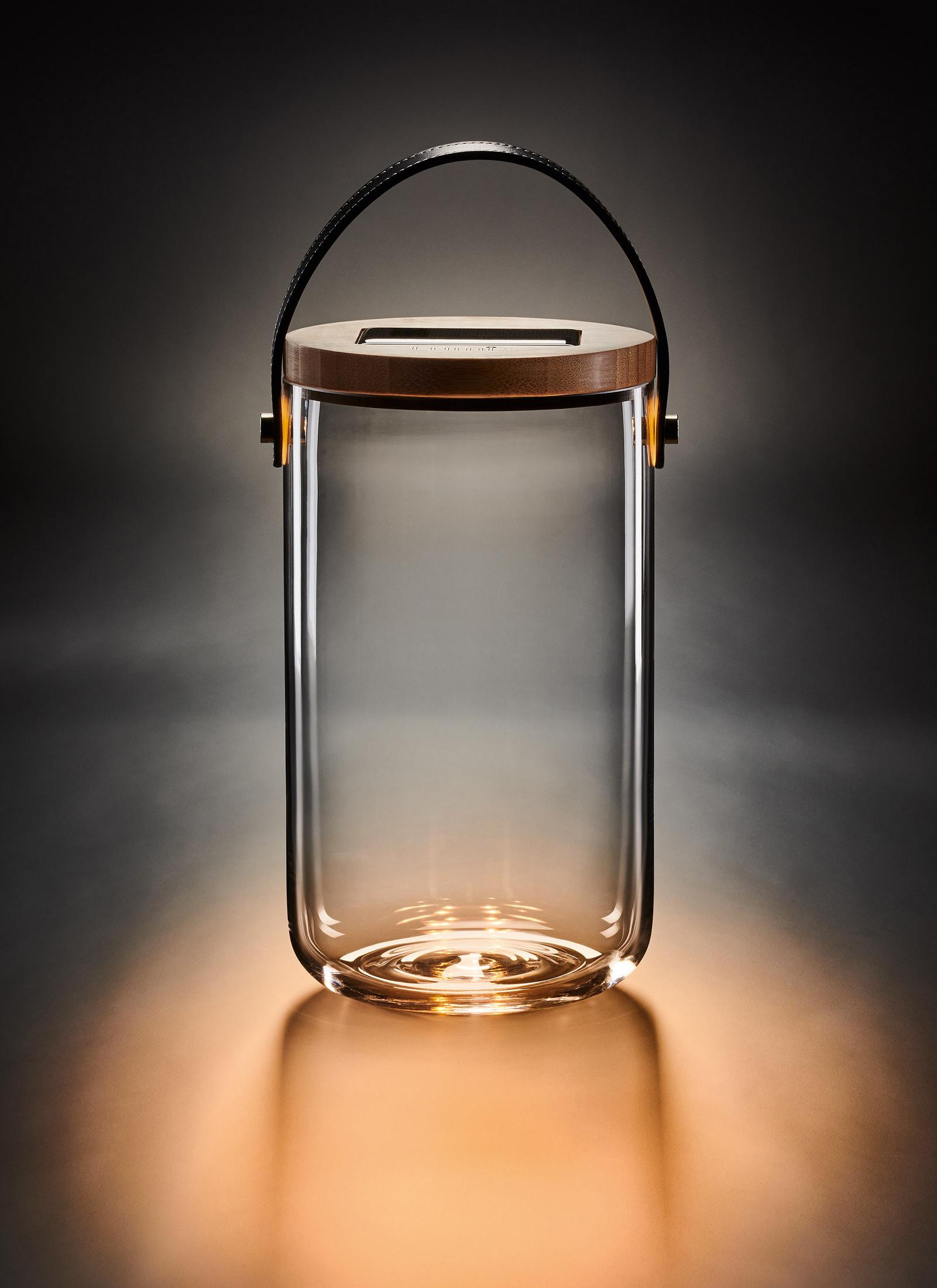 Eine klare Glaslaterne mit Holzdeckel und Ledergriff. Sie steht auf einer Oberfläche mit warmem Schein.