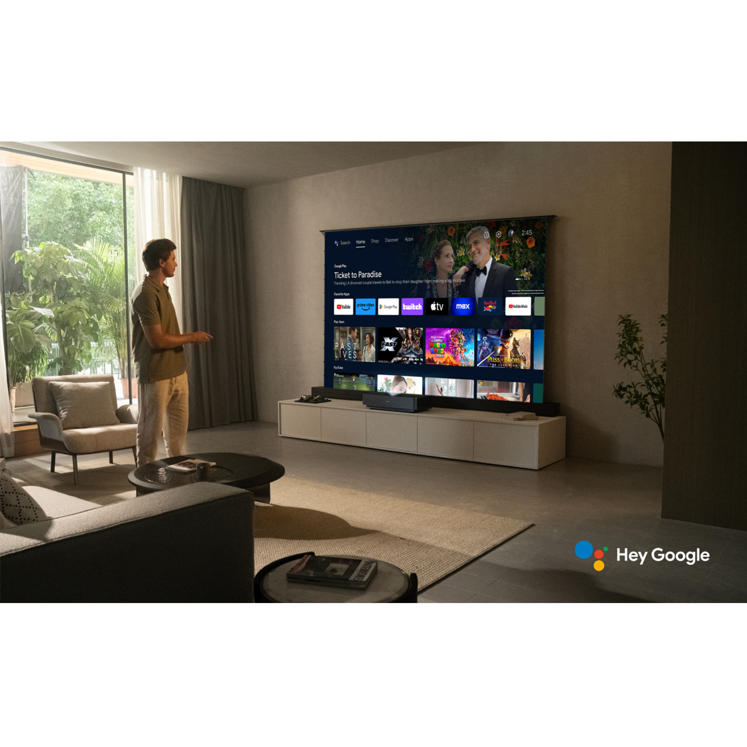 Mężczyzna stoi w salonie, trzymając pilota. Duży telewizor wyświetla aplikacje. W pokoju znajdują się kanapa, krzesła i dywan.