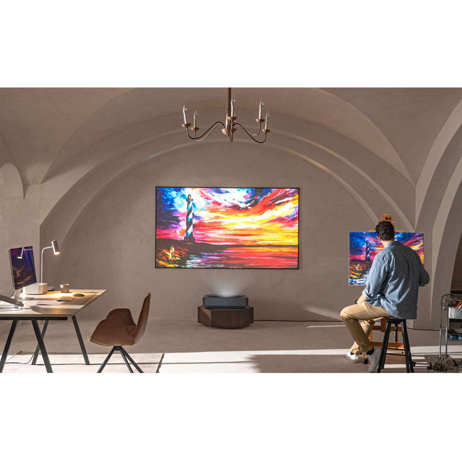 Mężczyzna maluje w pokoju z dużym ekranem wyświetlającym sztukę. Projektor stoi na drewnianej skrzyni. Widoczne są biurko i krzesło.