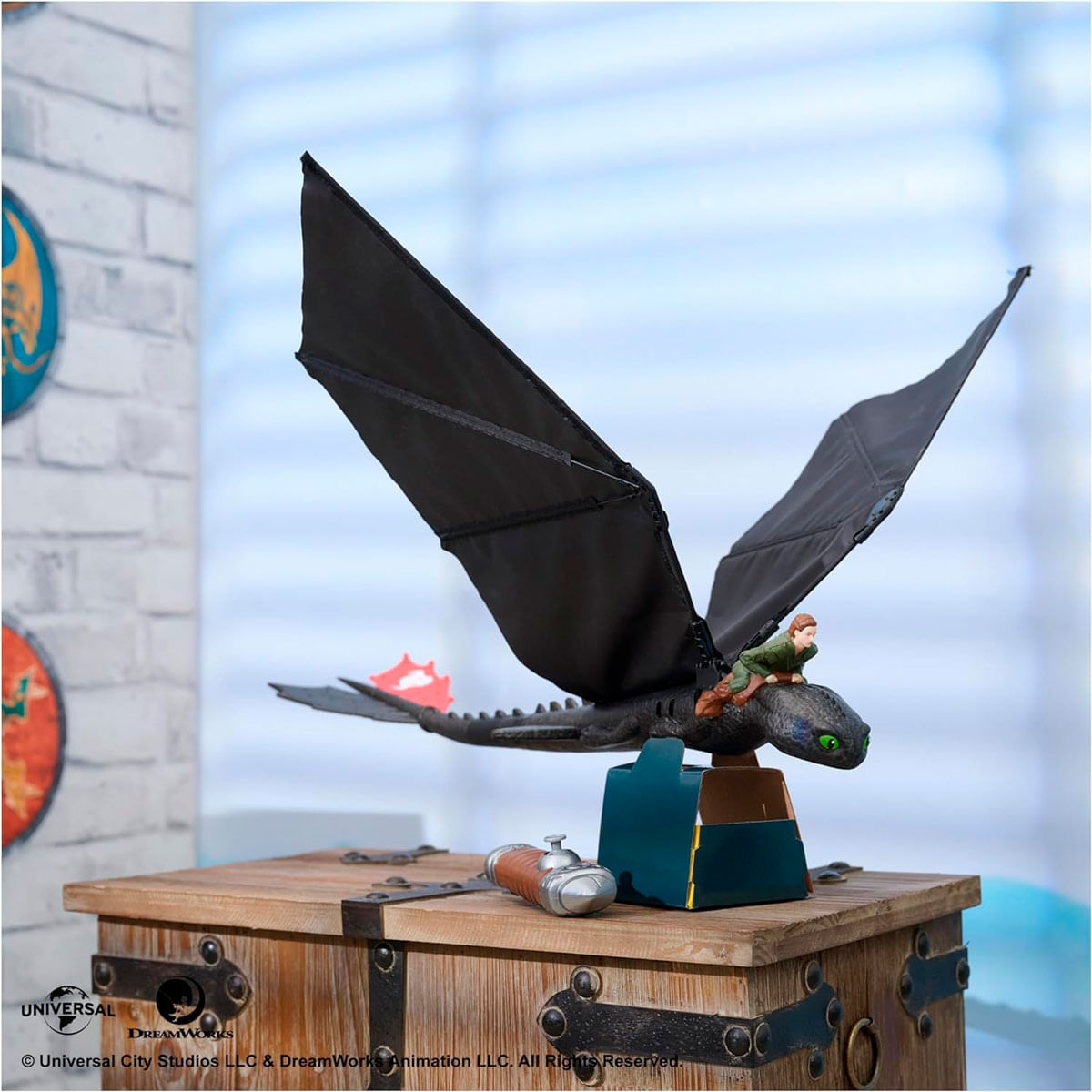 Un dragón de juguete con un jinete en una caja de madera, alas negras extendidas. Una pared de ladrillo y una ventana en el fondo.