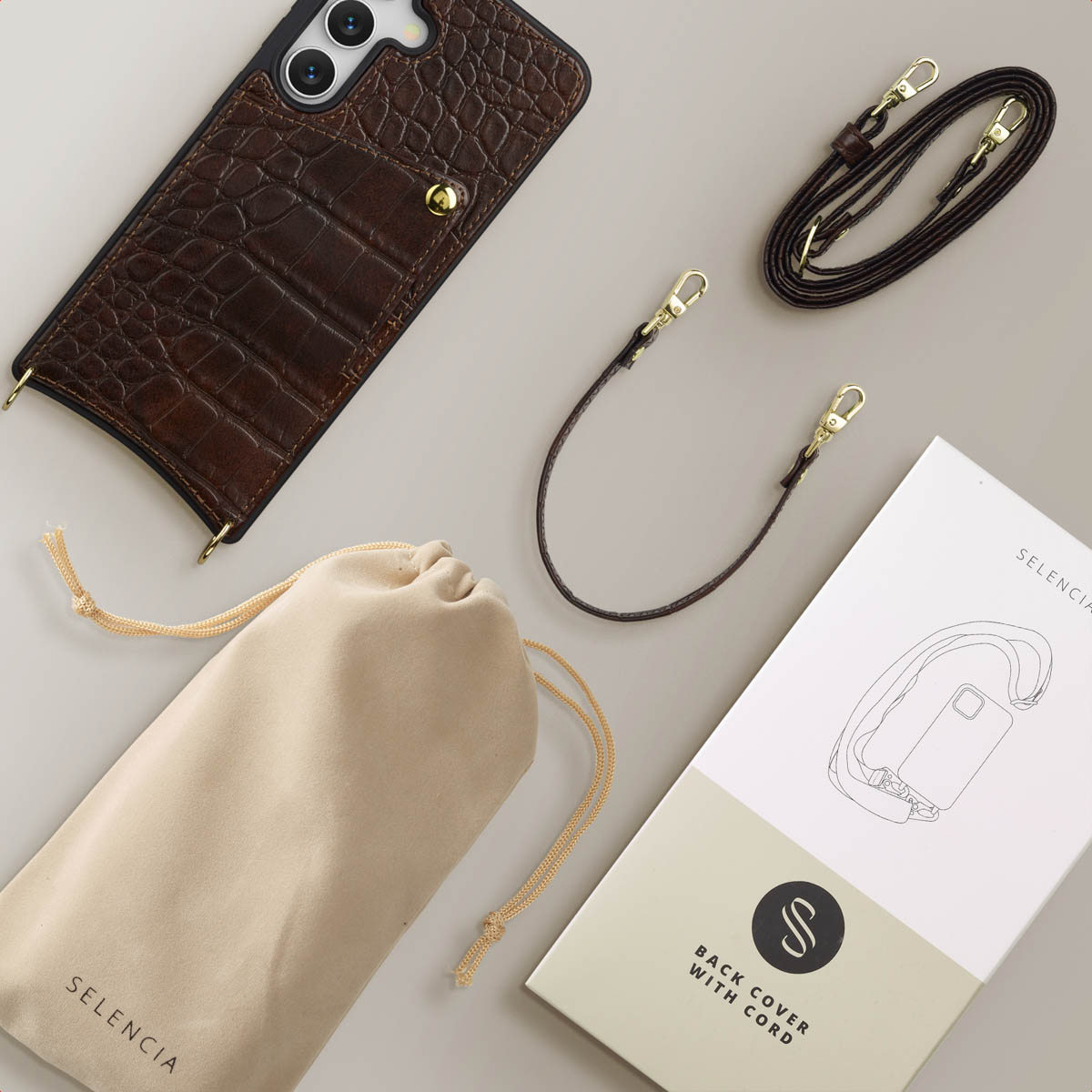 Bruine telefoonhoes met riem, een beige zakje, een boekje en een donkerbruine riem op een lichtgrijze ondergrond. Accessoires.
