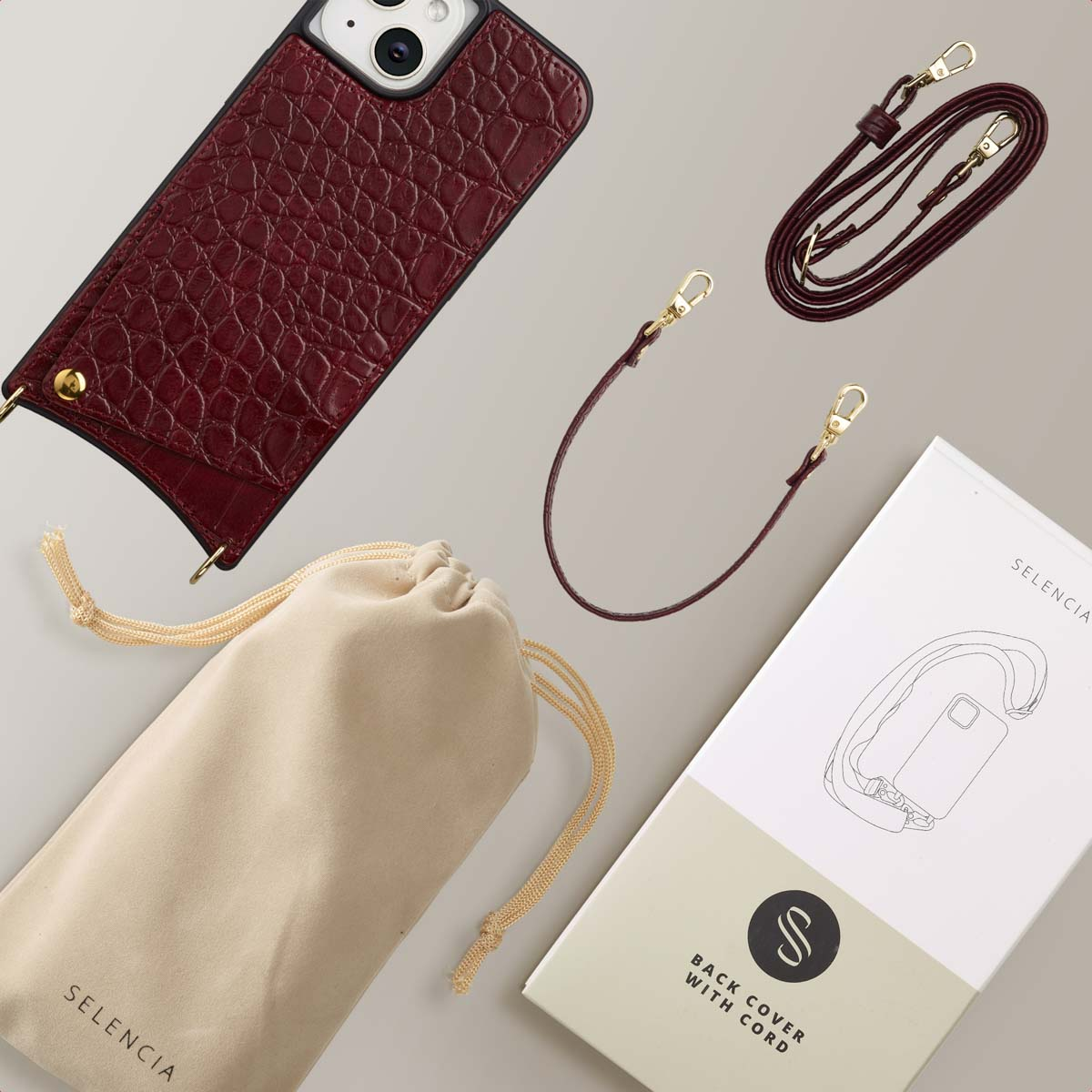 Een telefoonhoesje, riem, zakje en doos worden getoond. De case is bordeauxrood, met een riem en een zakje. De doos toont een telefoon met een riem.