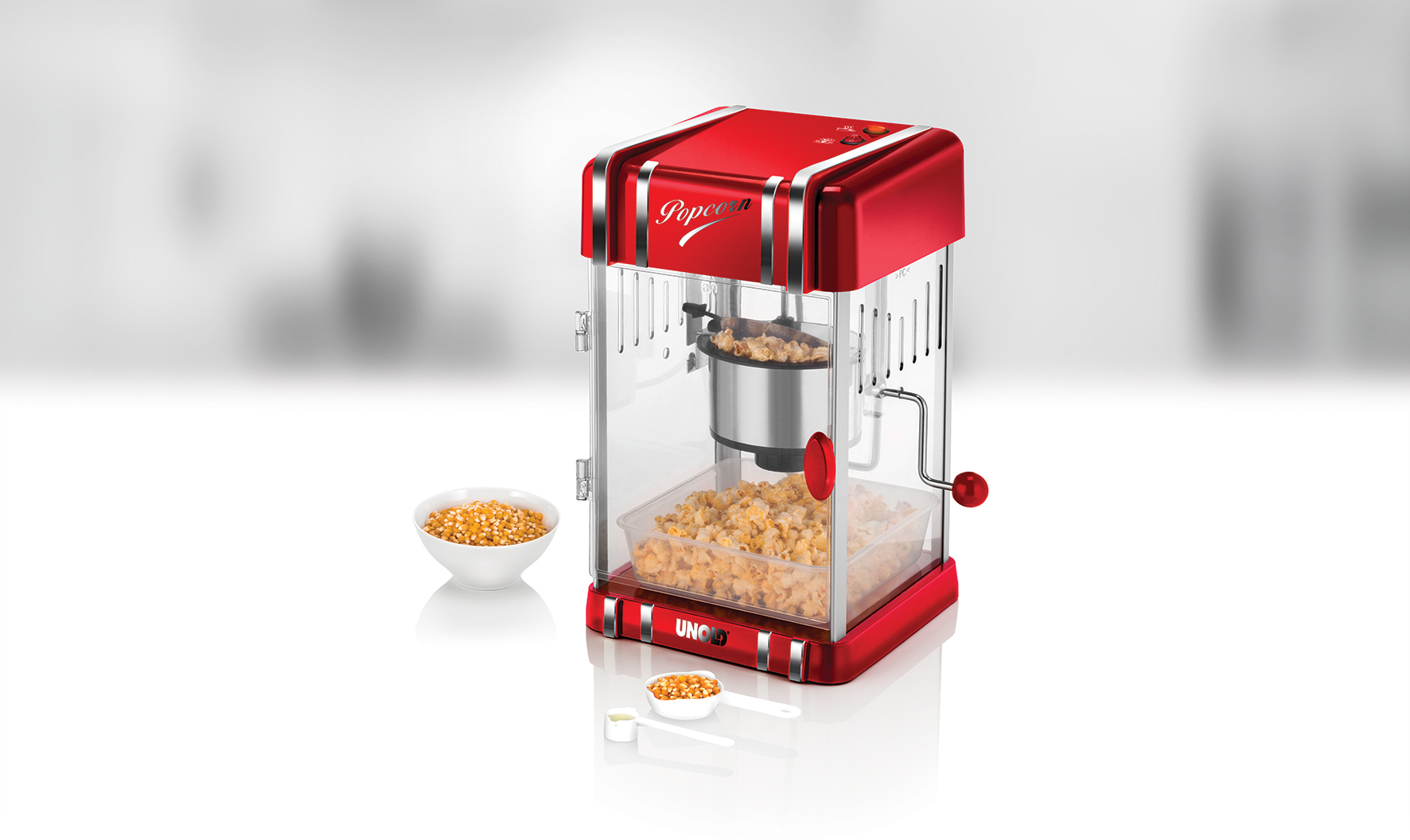 Ein roter Popcorn-Maker mit transparenten Seiten, mit einer Schüssel mit Maiskörnern und Popcorn.