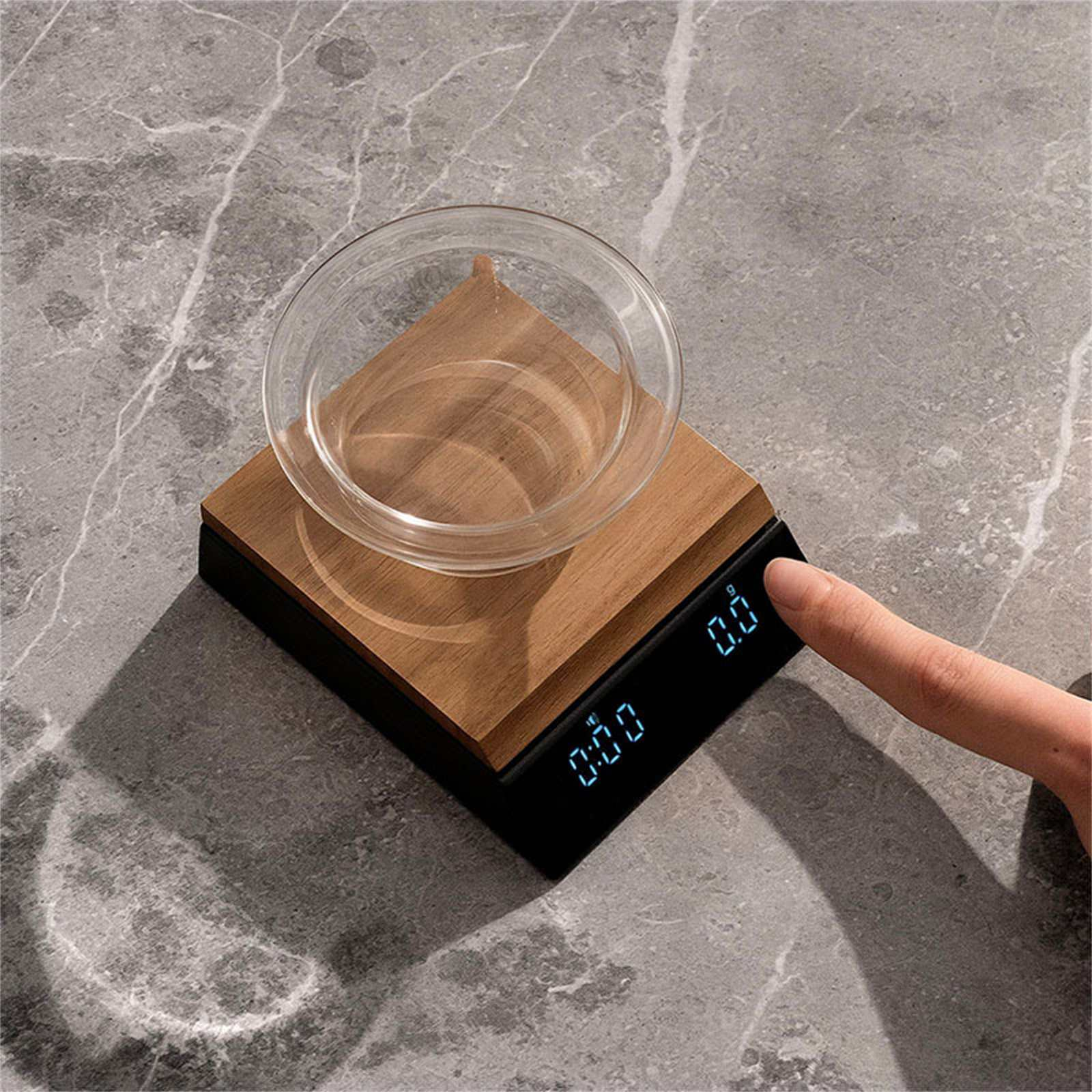 Eine Waage mit Holzaufsatz und Glasschale. Ein Finger berührt die Waage. Der Hintergrund ist ein Steintisch.