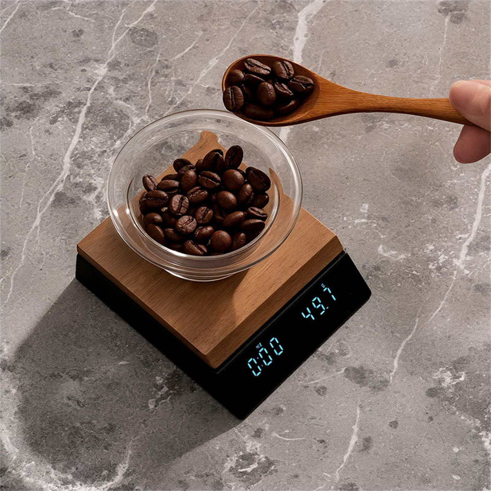 Kaffeebohnen in einer Glasschale auf einer Waage. Ein Holzlöffel hält mehr Bohnen. Grauer Marmorhintergrund.